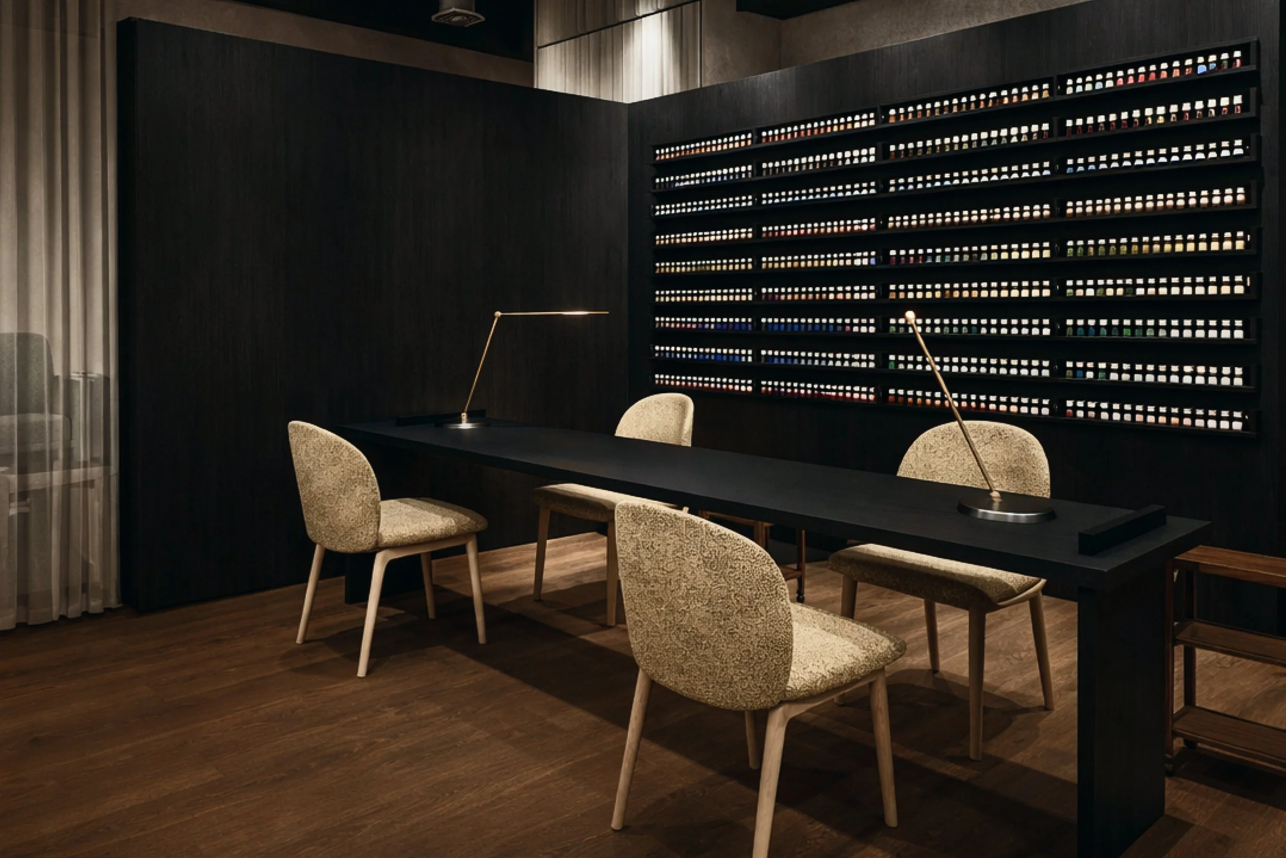 A modern, dark-themed conference room with a long black table, four beige upholstered chairs, a wall display of small colorful items, and two sleek brass desk lamps.