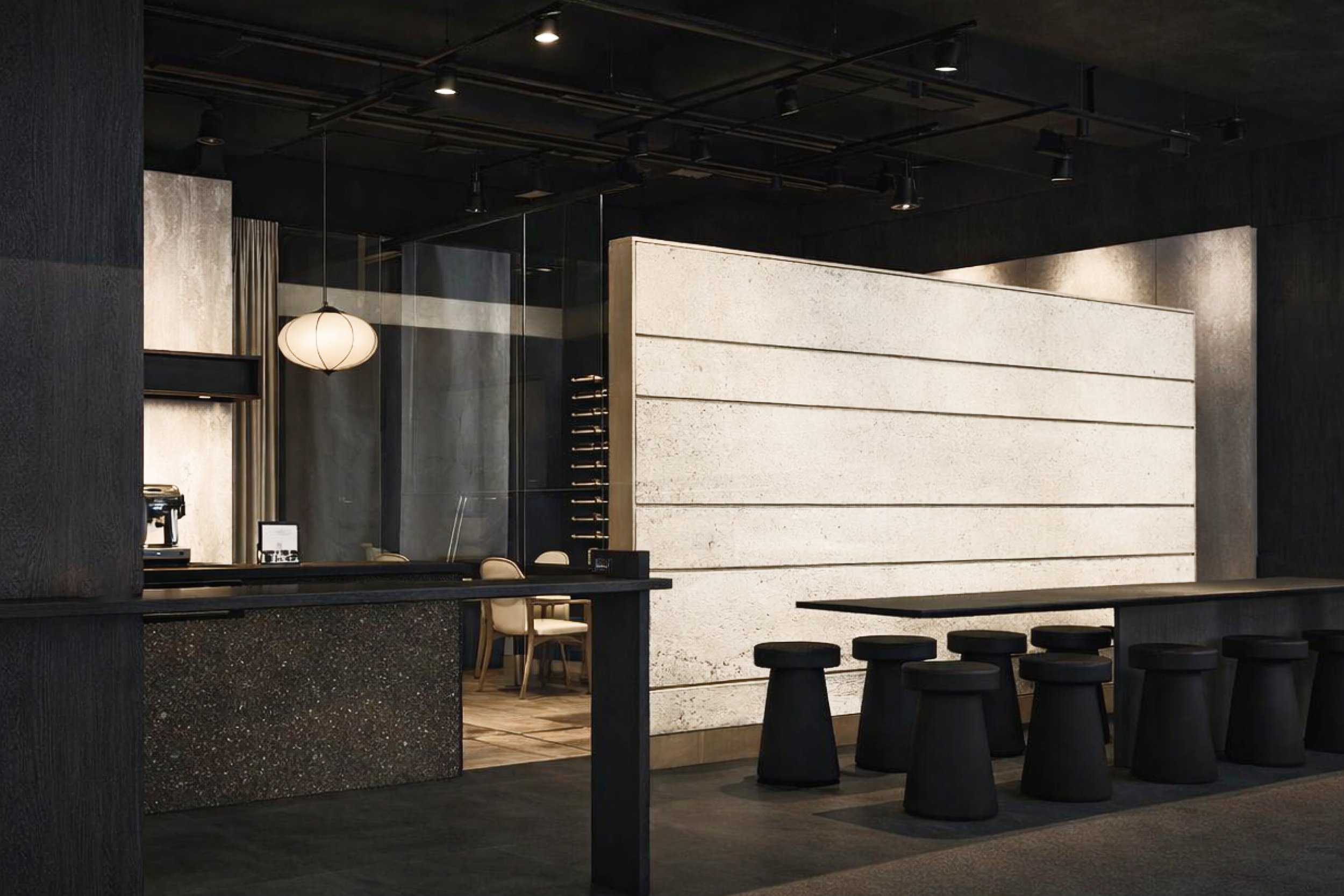 Modern interior of a cafe or restaurant with black and white decor, stools at a long table, chairs, pendant lighting, and a counter with a coffee machine.