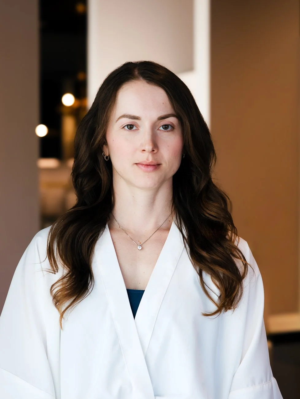 A woman with long dark wavy hair, wearing a white coat, standing in an indoor setting with soft lighting.