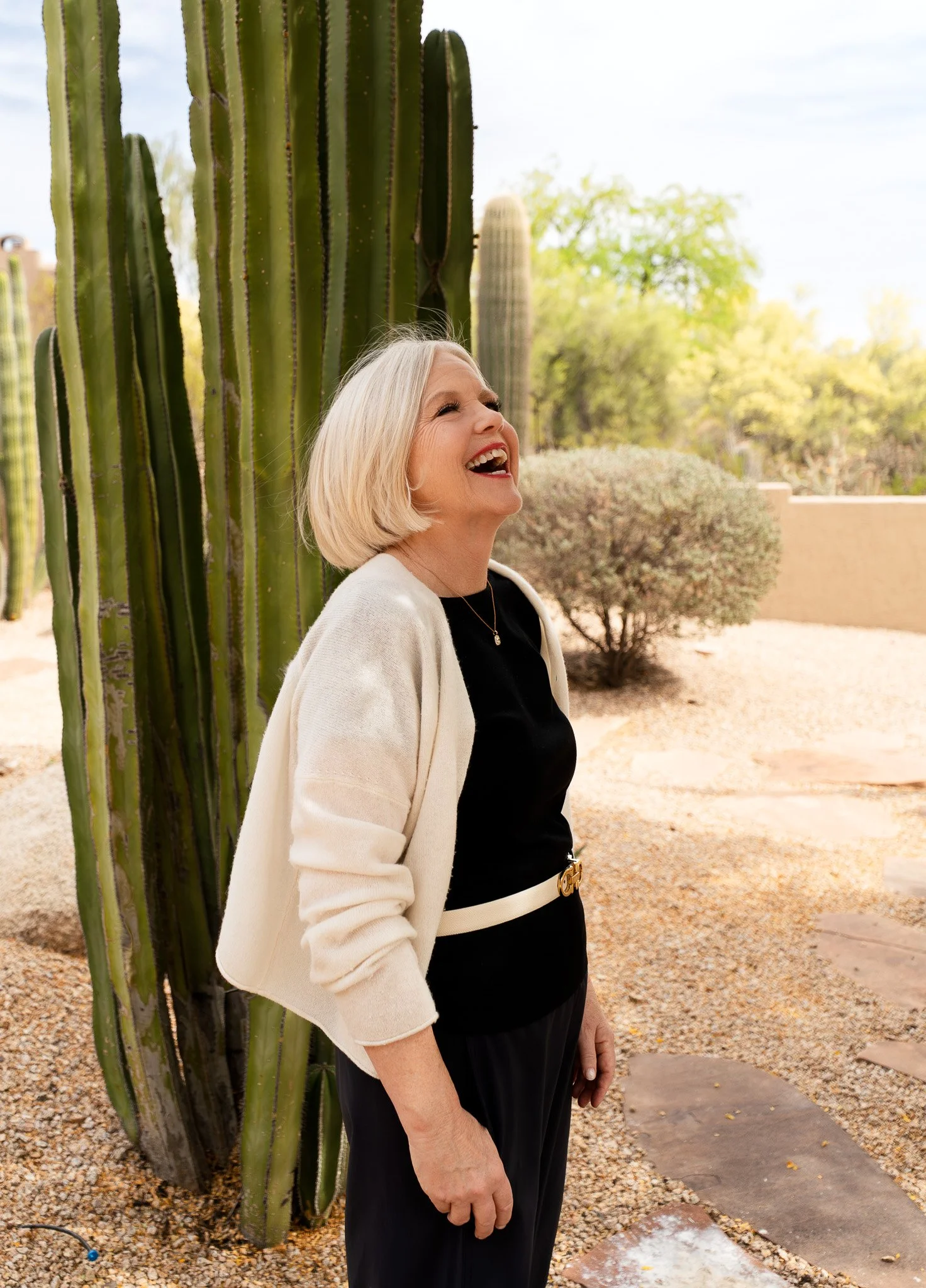 Woman with short blonde hair with a black dress and white sweater laughing standing in her backyard in Arizona with a large green cactus