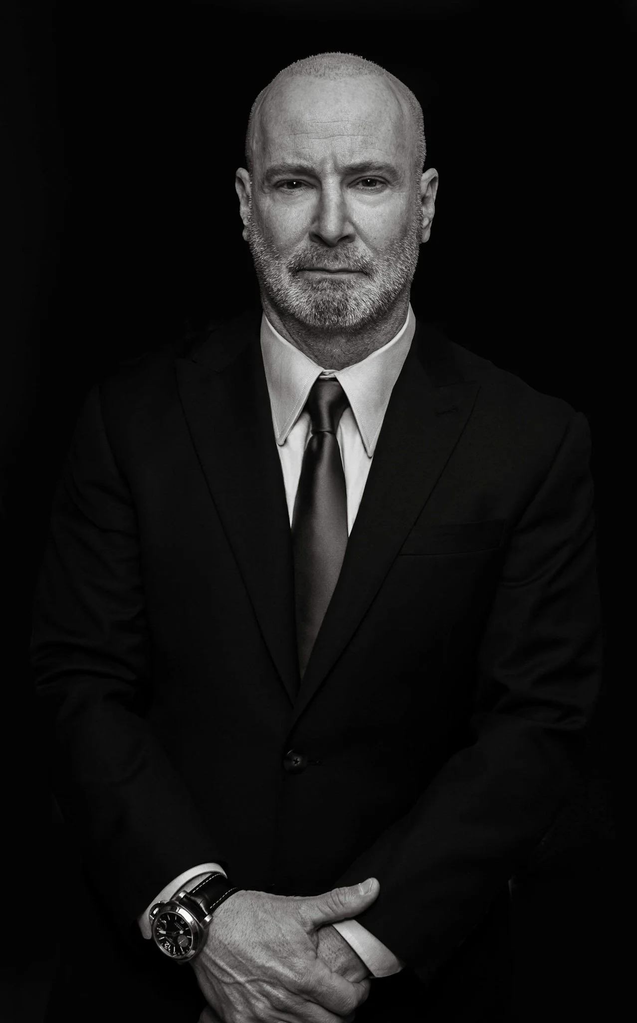 Black and white portrait of a middle-aged man with a beard, wearing a dark suit, white shirt, and a dark tie, with his hands clasped in front of him against a black background.