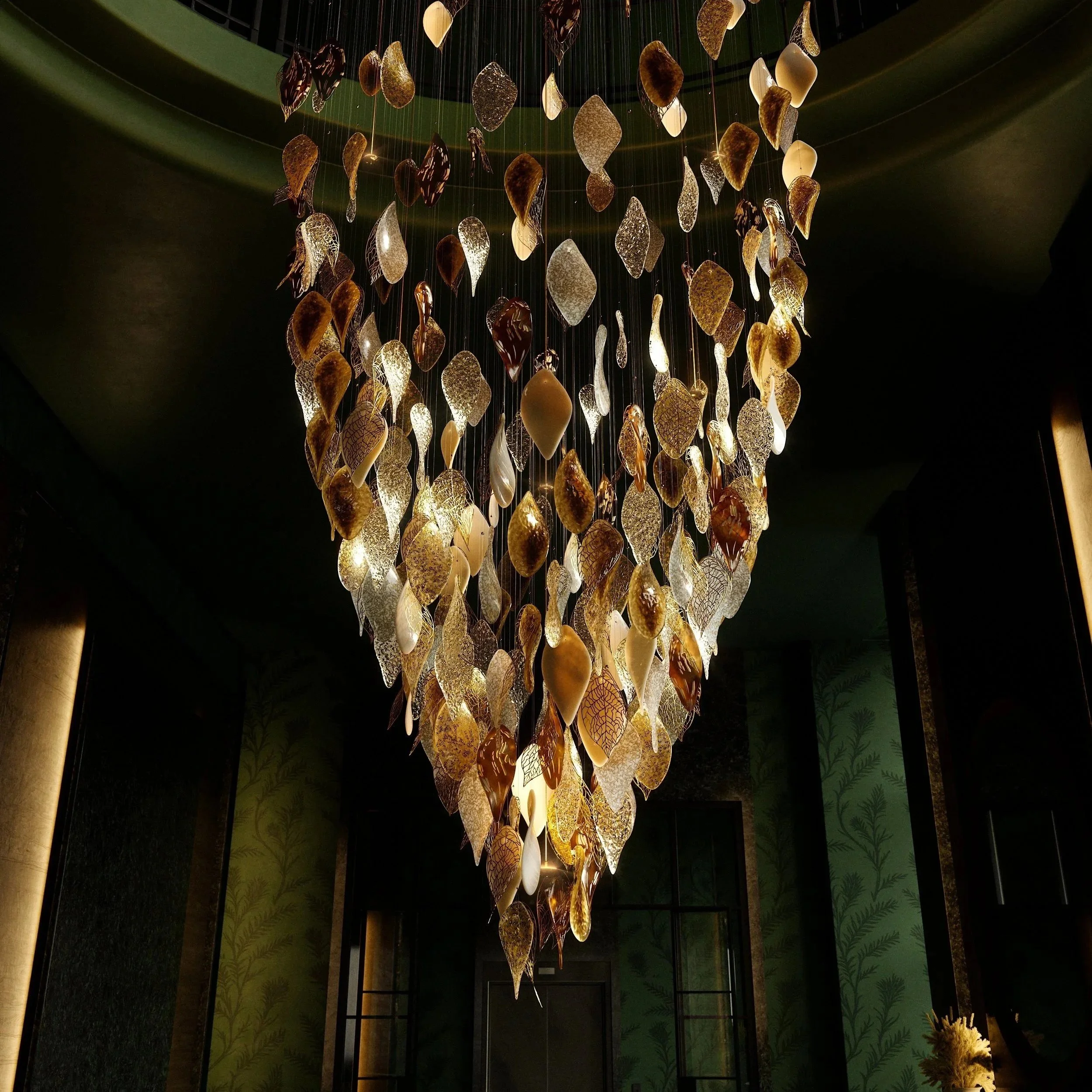 Elegant chandelier of cascading glass leaves in amber, gold, and clear tones, suspended on wires, glowing warmly and forming a sculptural, organic chandelier of light.
