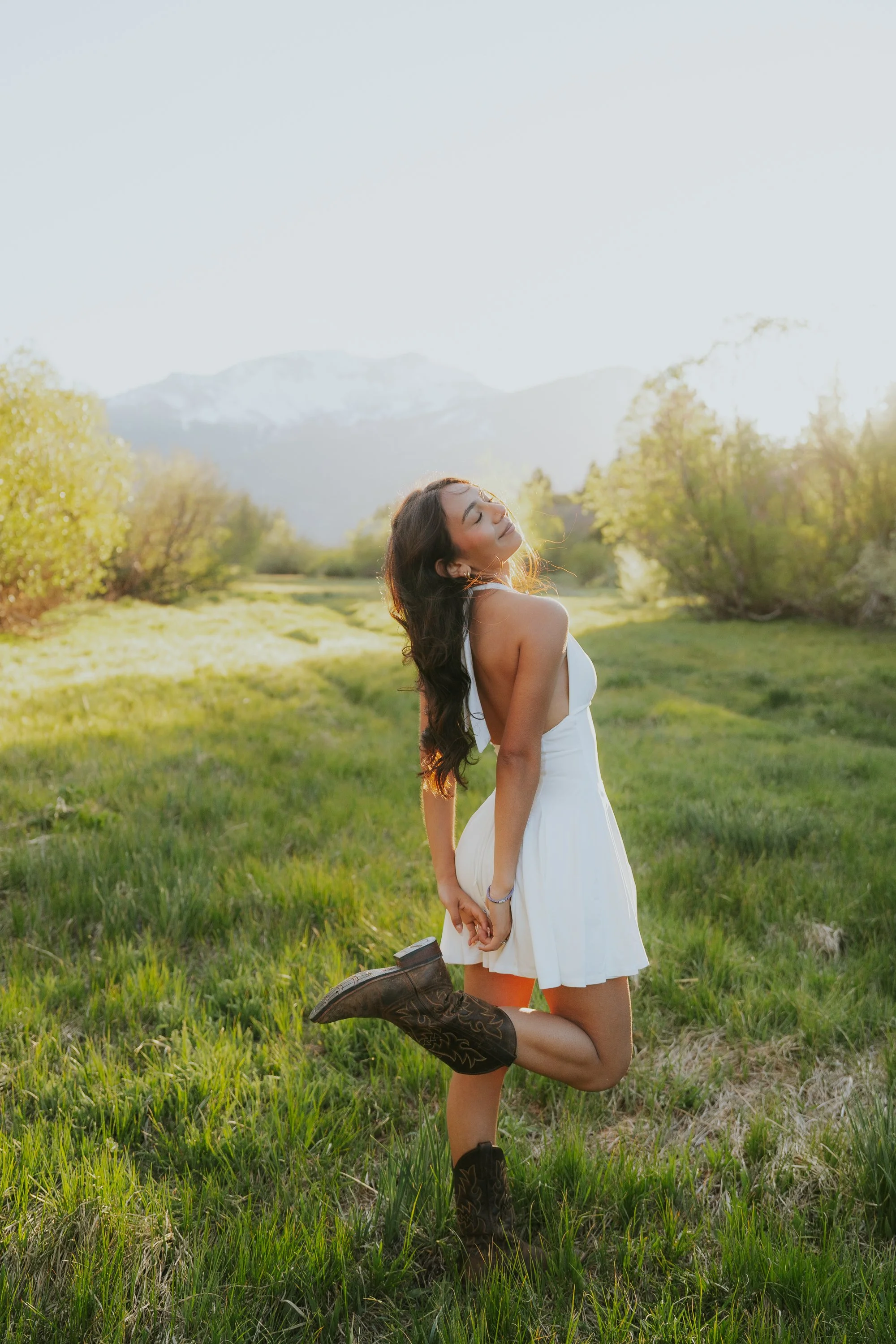 A woman in a white dress and cowboy boots kneels on green grass in a scenic outdoor setting with trees and mountains in the background, smiling with her eyes closed.