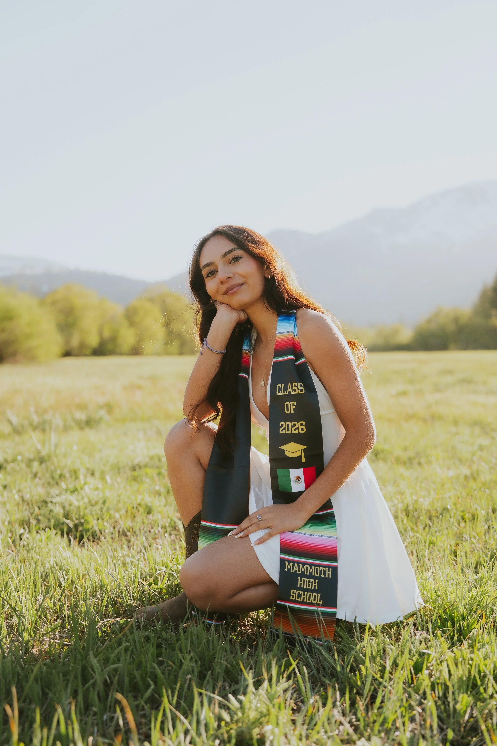 Young woman kneeling on grass outdoors wearing a white dress and a graduation stole that reads "Mammoth High School" and "Class of 2026" with a Mexican flag. Mountains and trees are in the background.