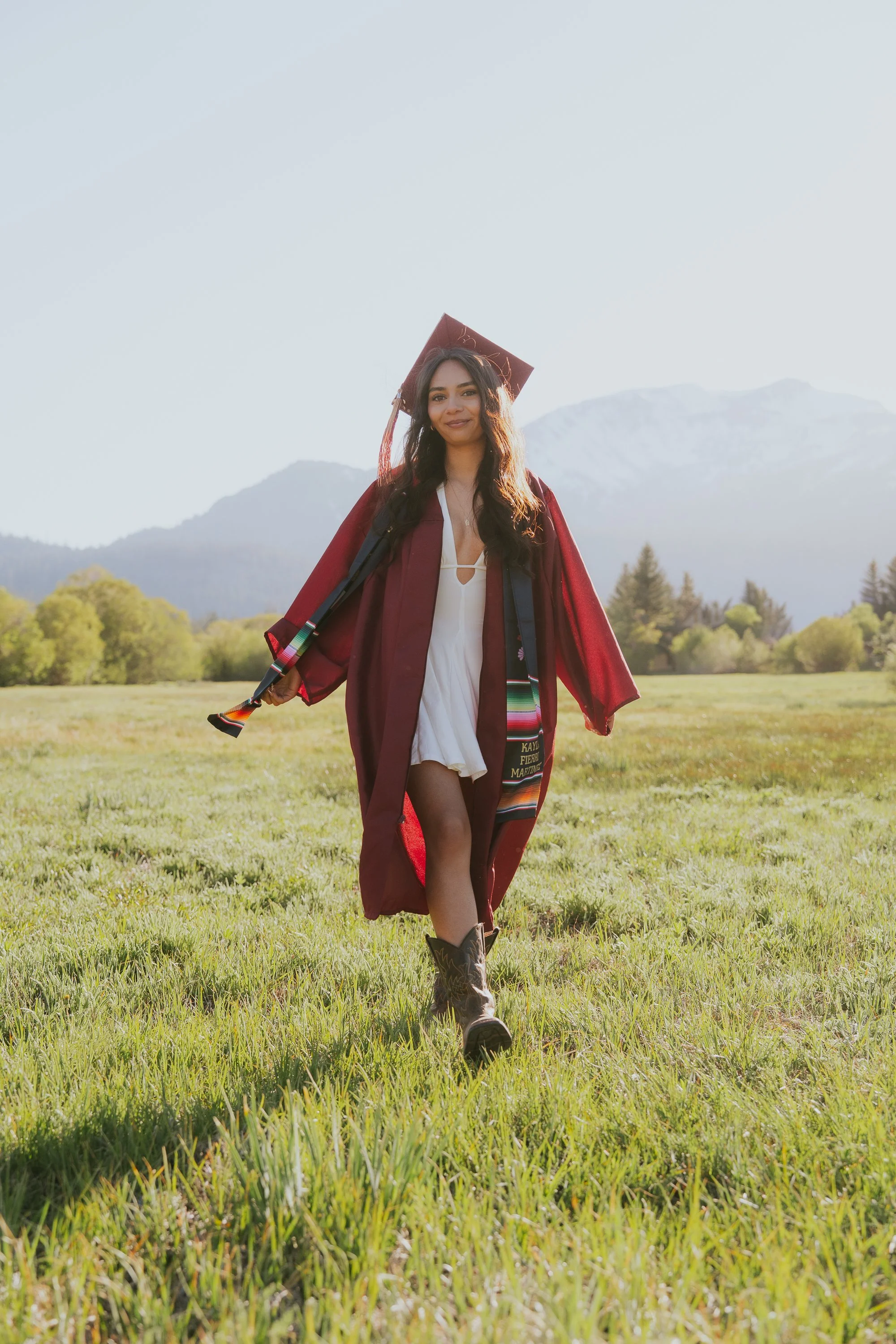 A young woman in a burgundy graduation cap and gown walking across a green field with mountains in the background, smiling at the camera.