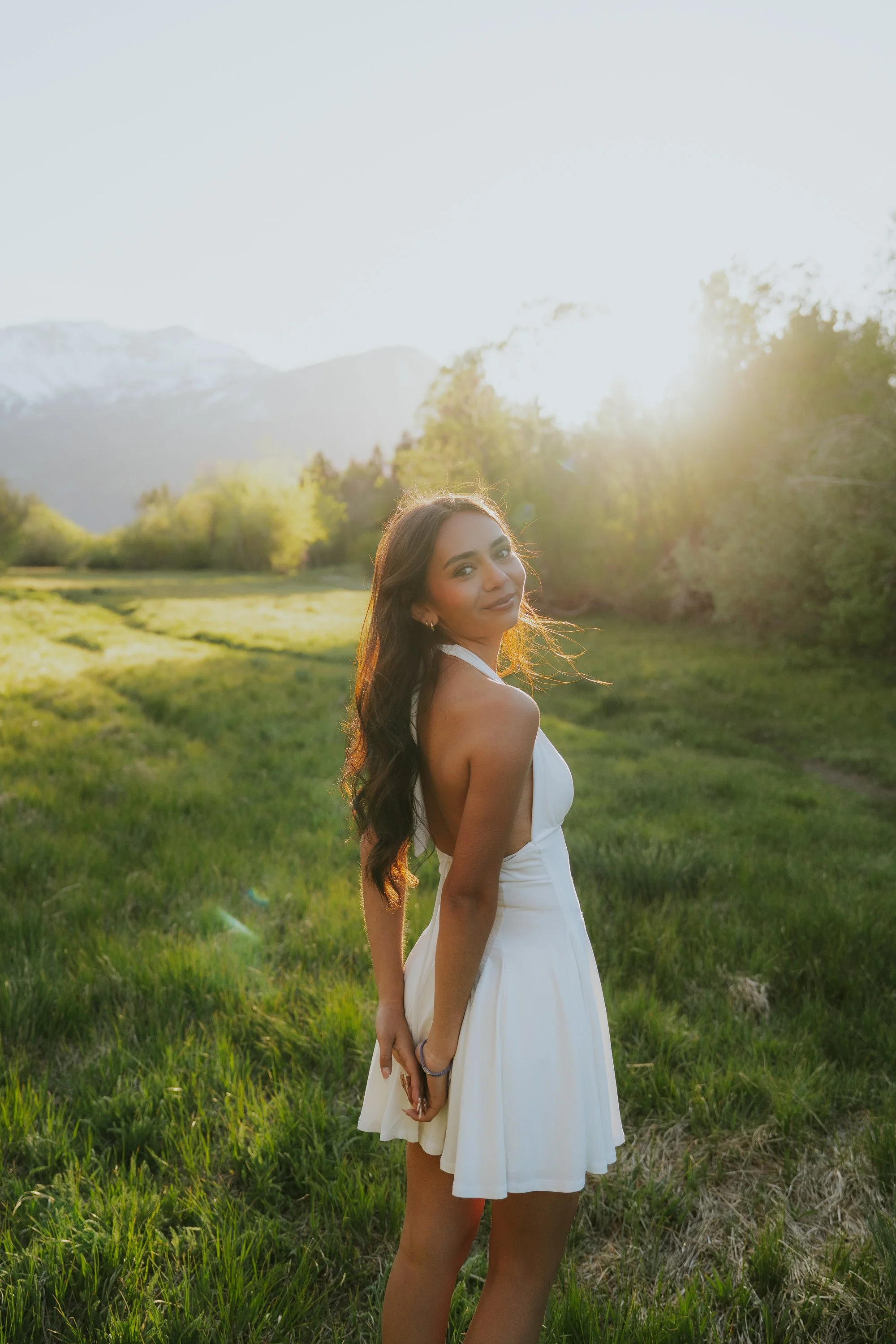 A young woman with long brown hair wearing a white dress standing in a lush green field during sunset with mountains in the background.