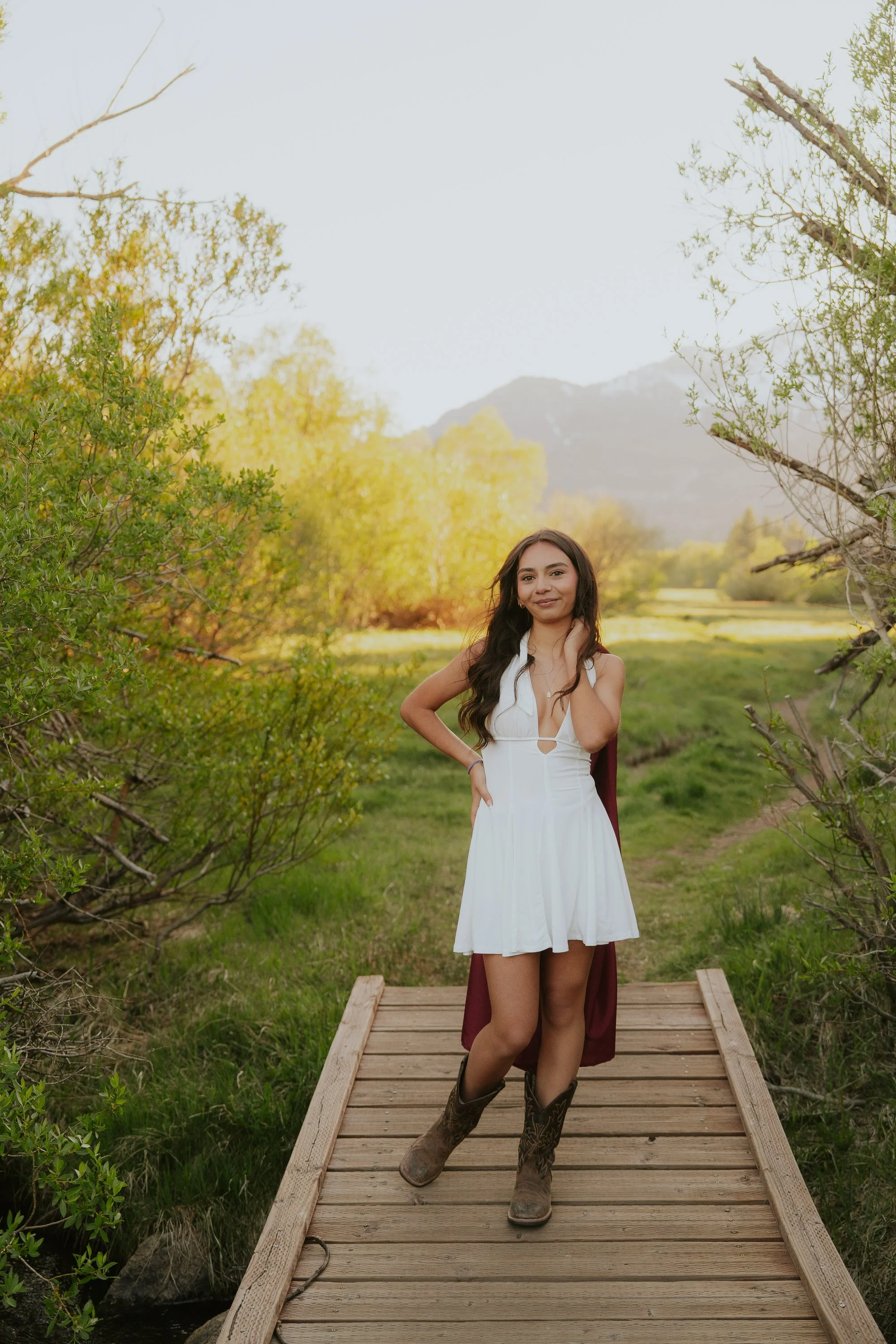 A woman standing on a wooden footbridge in a lush, green outdoor setting with trees and mountains in the background, wearing a white dress and cowboy boots.