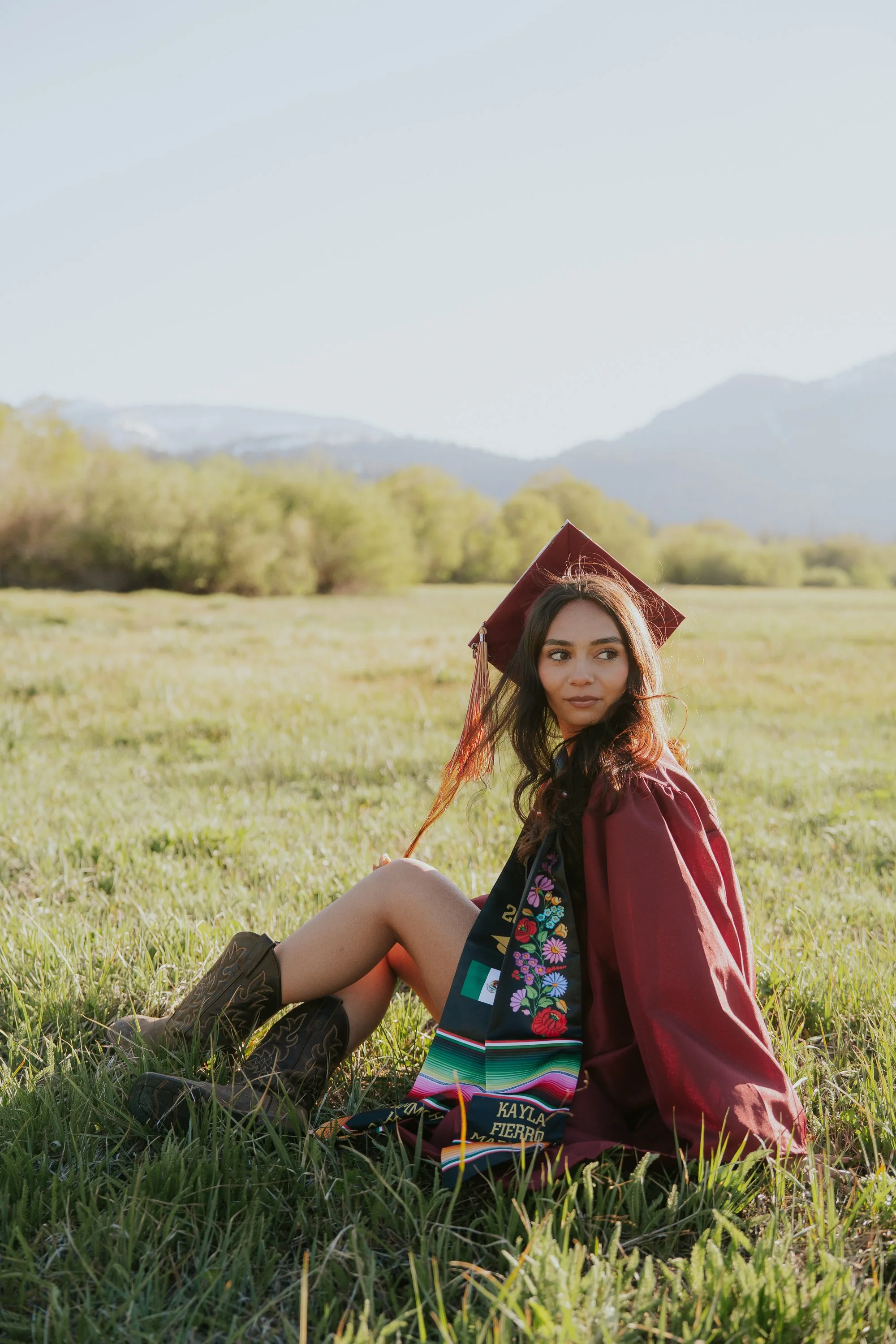 A young woman wearing a maroon graduation gown and cap sits on green grass in a field, with mountains and trees in the background. She has black cowboy boots and a colorful sash that says 'Kayla Fierro.'