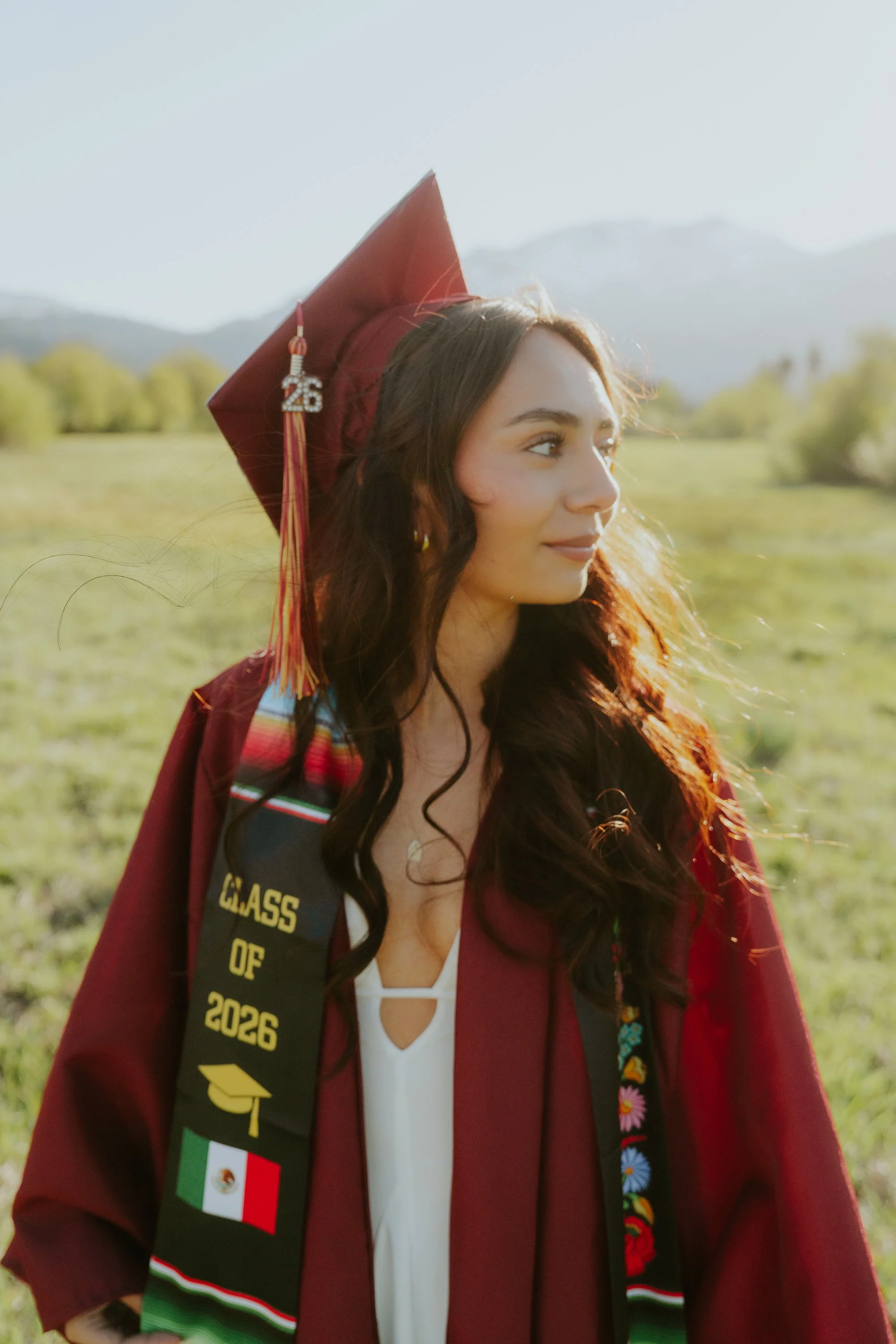 Young woman in a maroon graduation gown and cap, outdoors in a field with mountains in the background, smiling subtly.
