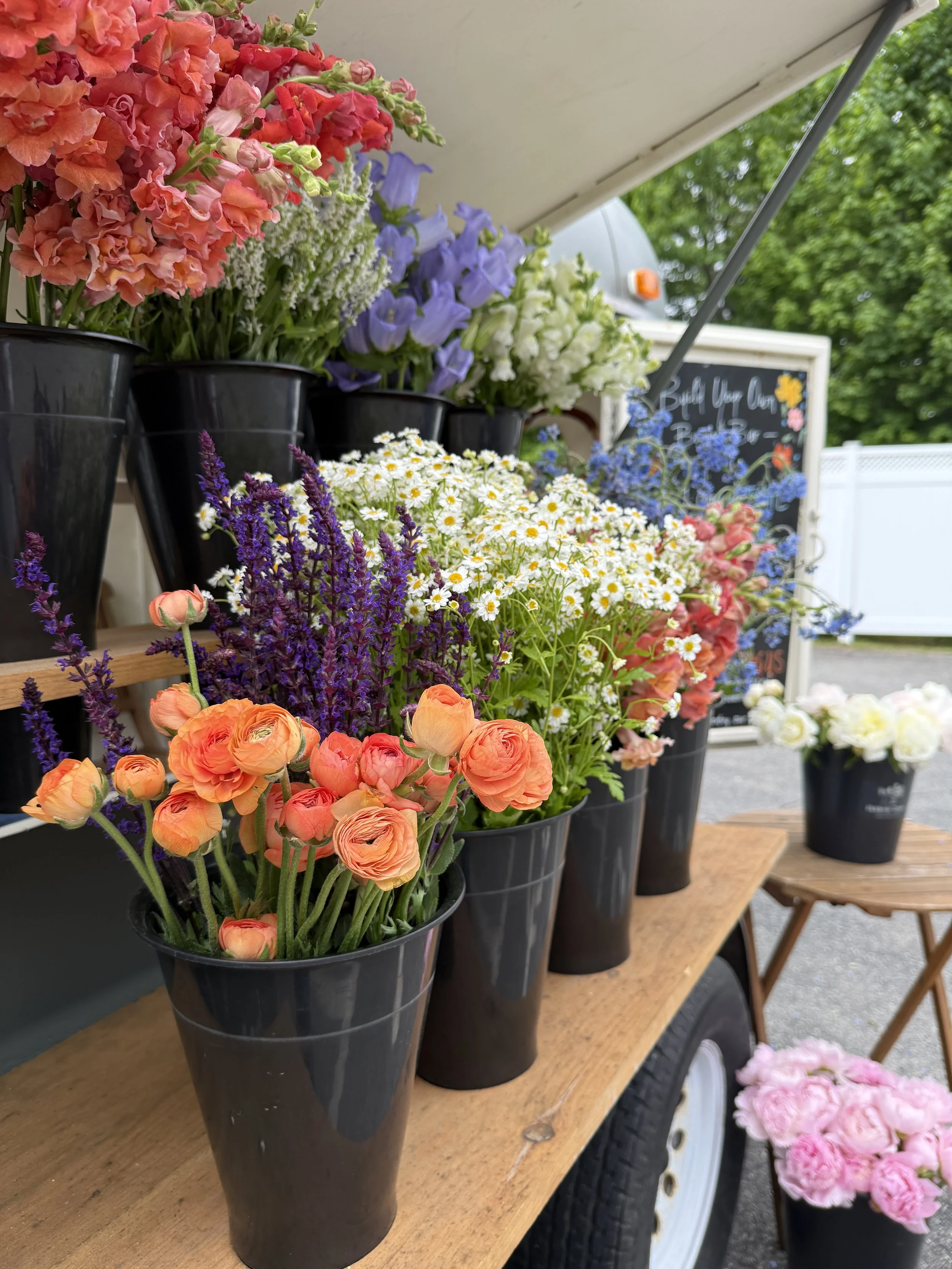 Various colorful flowers in black pots displayed on a street vendor's stand, including orange ranunculus, purple lavender, white daisies, blue geraniums, and peach snapdragons, with a chalkboard sign in the background.
