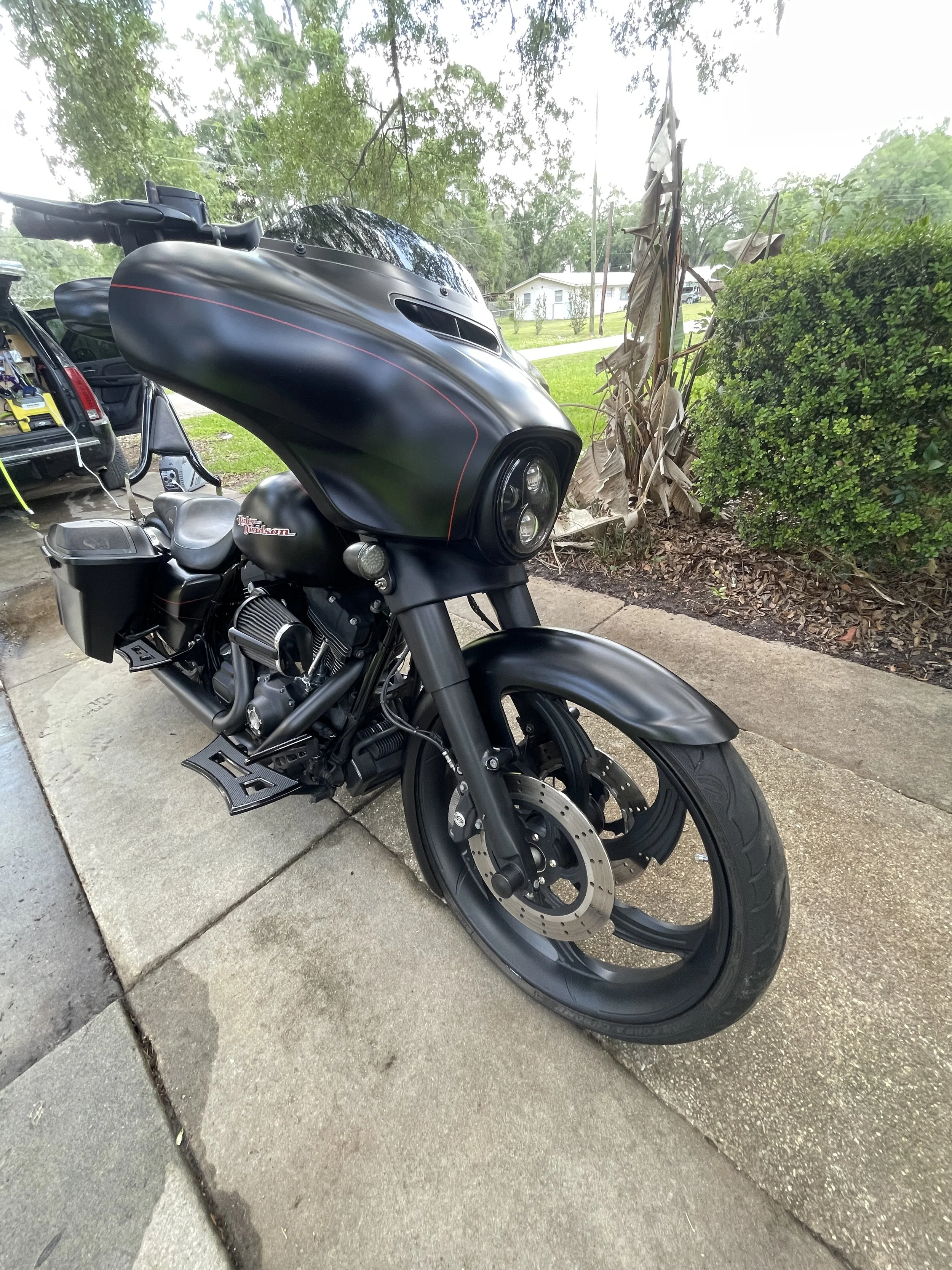 A black Harley Davidson motorcycle parked on a residential sidewalk with a house and trees in the background.