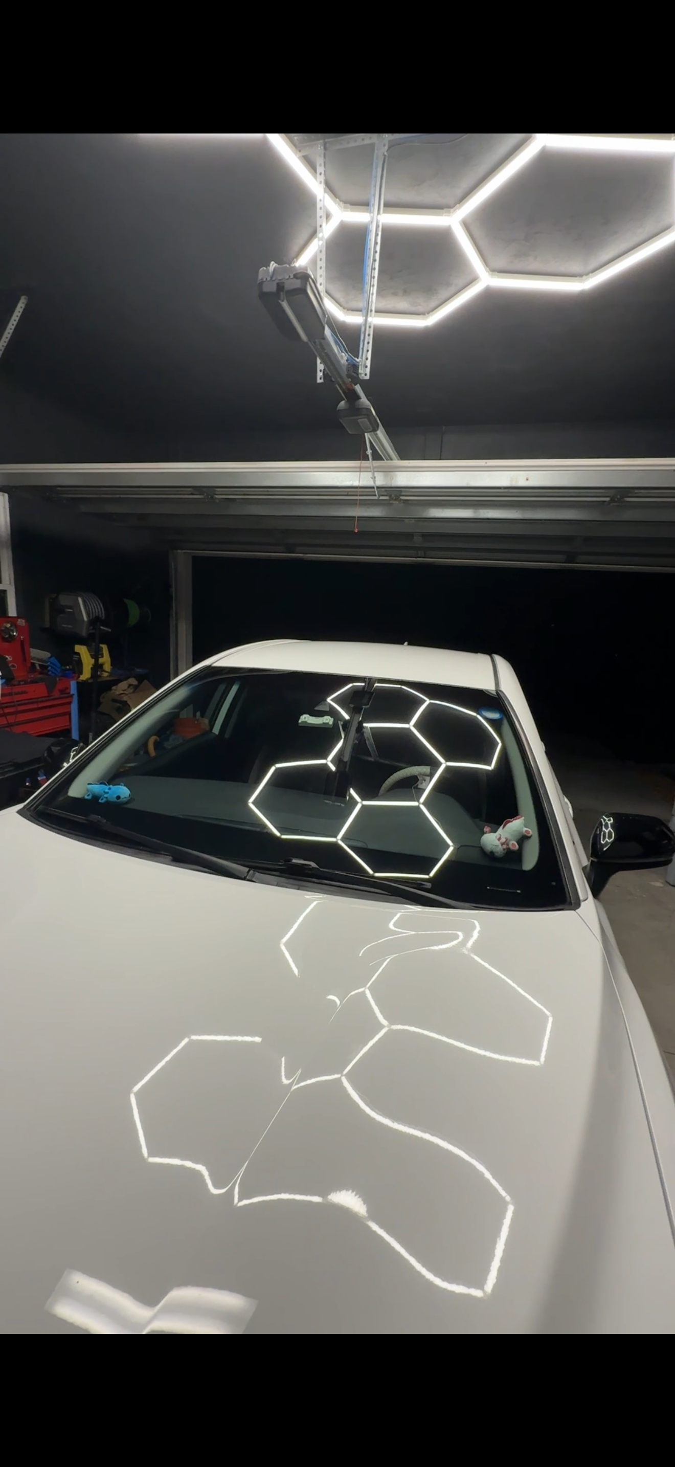 A white car parked in a garage with hexagon-shaped LED lights reflected on its hood and windshield. The garage has black walls and various tools and equipment visible in the background.