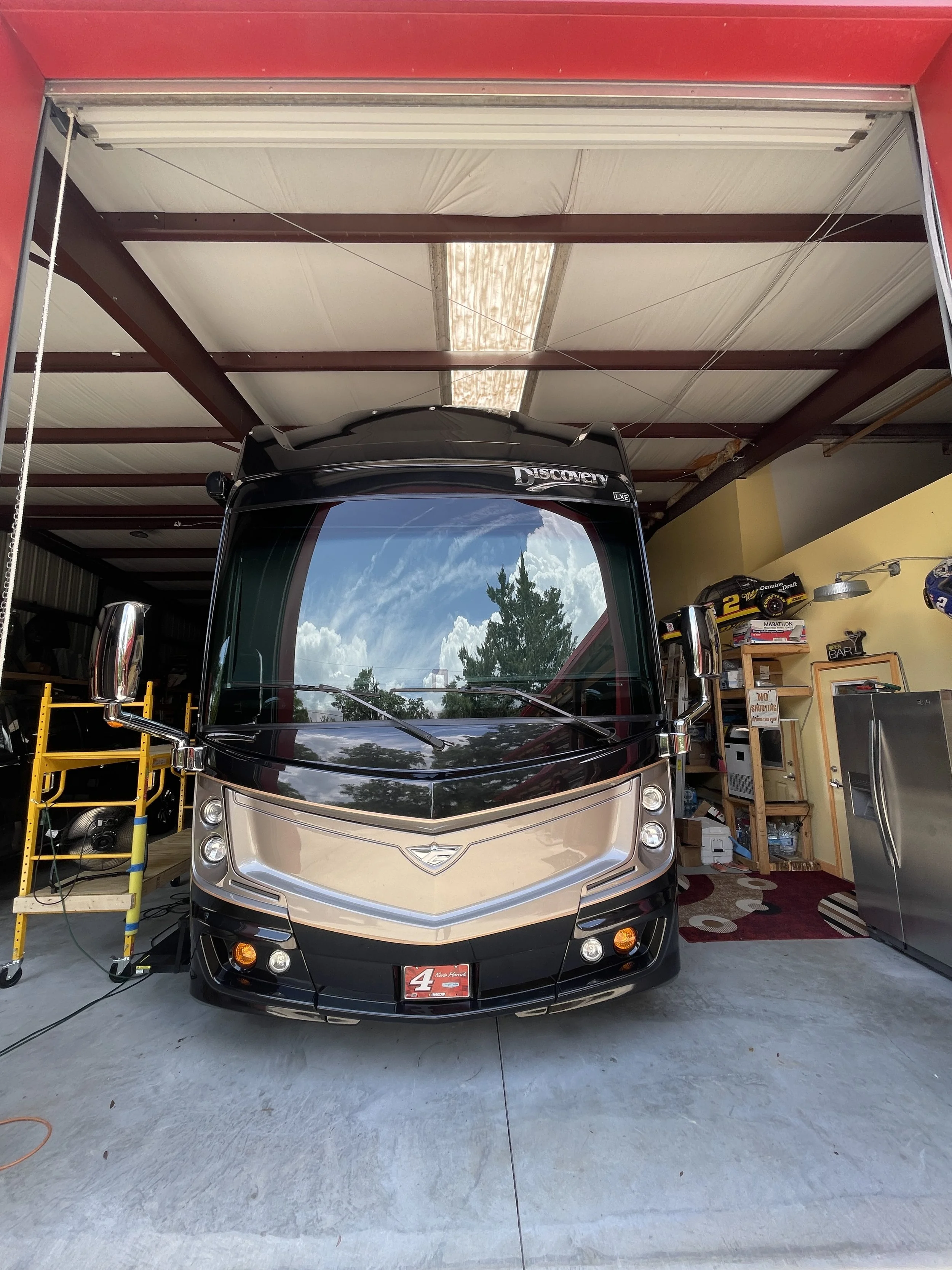 Front view of a black and beige RV parked inside a garage, with a reflection of clouds and trees on the windshield. The garage has shelves with various items, including books, boxes, and a small model race car. A yellow toolbox is on the left side.