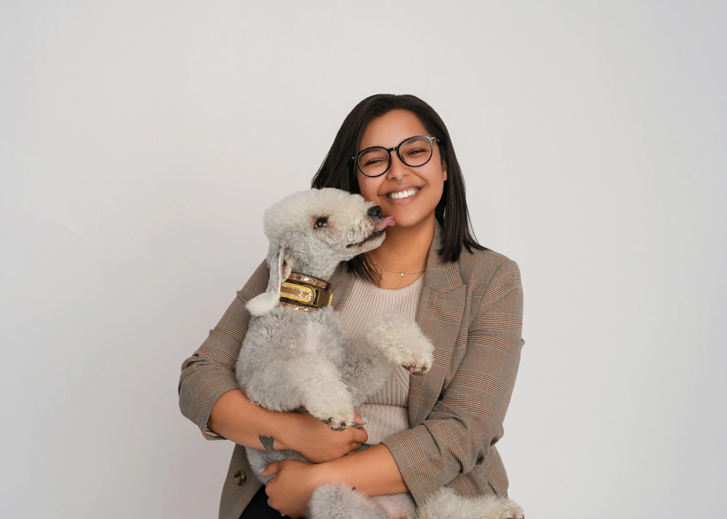 A woman with dark hair, glasses, and a plaid blazer is smiling while holding a Bedlington Terrier which is licking her face.