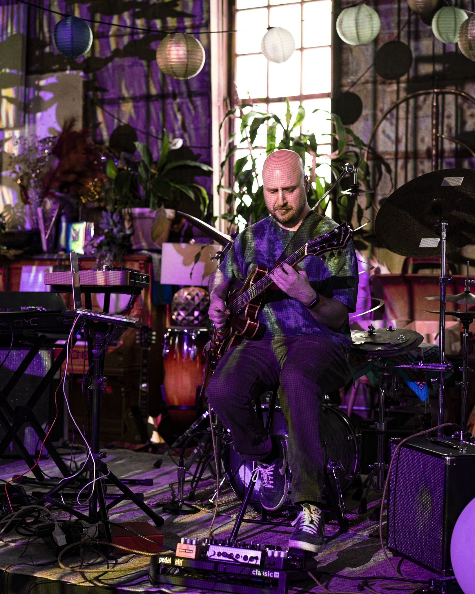 A man playing an acoustic guitar in a decorated indoor setting with plants, hanging paper lanterns, and a large window in the background.