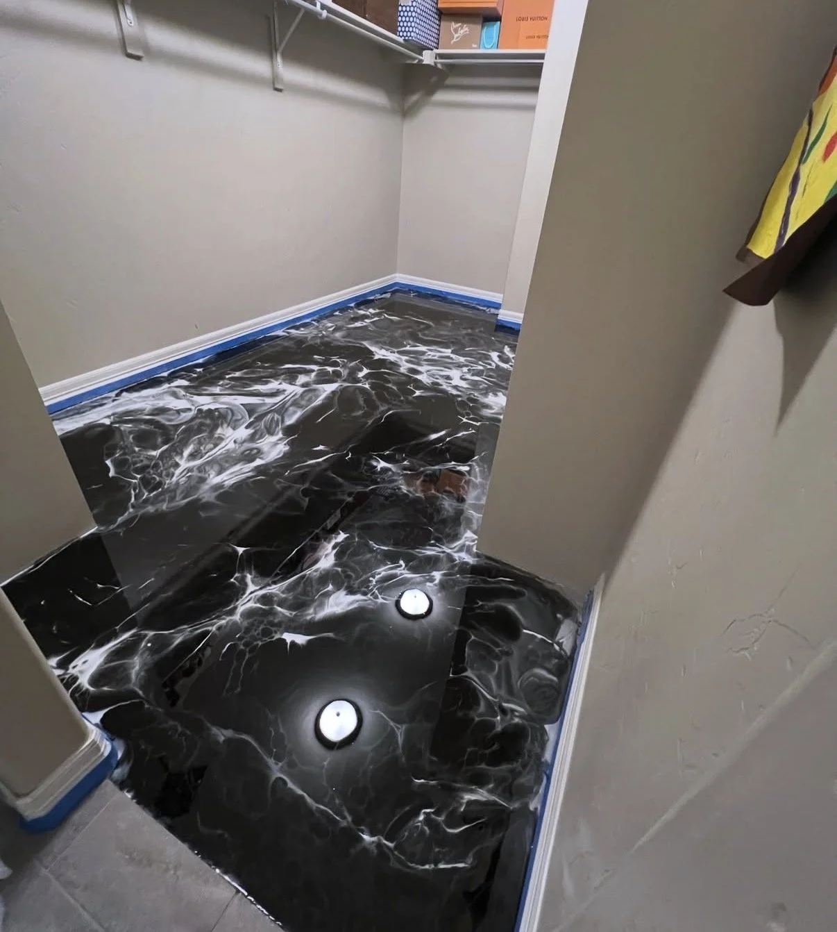 A bathroom with marble black and white flooring, with lights on the ceiling and beige walls. There are shelves above with boxes and small items.