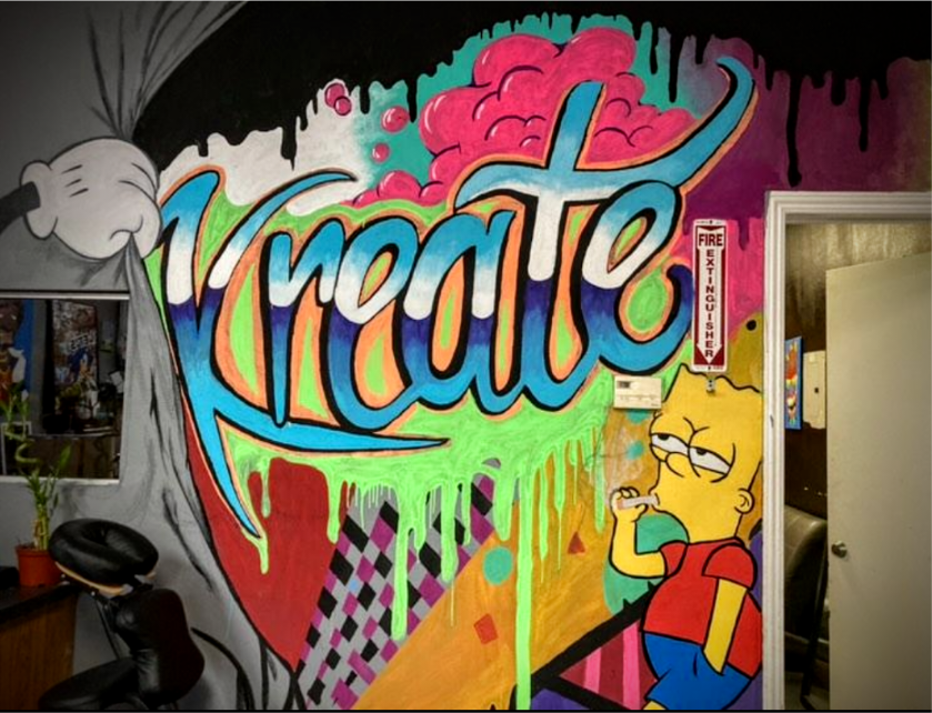 Colorful graffiti mural on a wall featuring the word 'Kraze' in bright, bold letters with drips above it. To the right, there is a cartoon drawing of Lisa Simpson from The Simpsons, with a bored or unimpressed expression, wearing a red shirt and blue shorts. The mural has vibrant colors including pink, green, and orange, and includes a small fire extinguisher sign and some abstract patterns.
