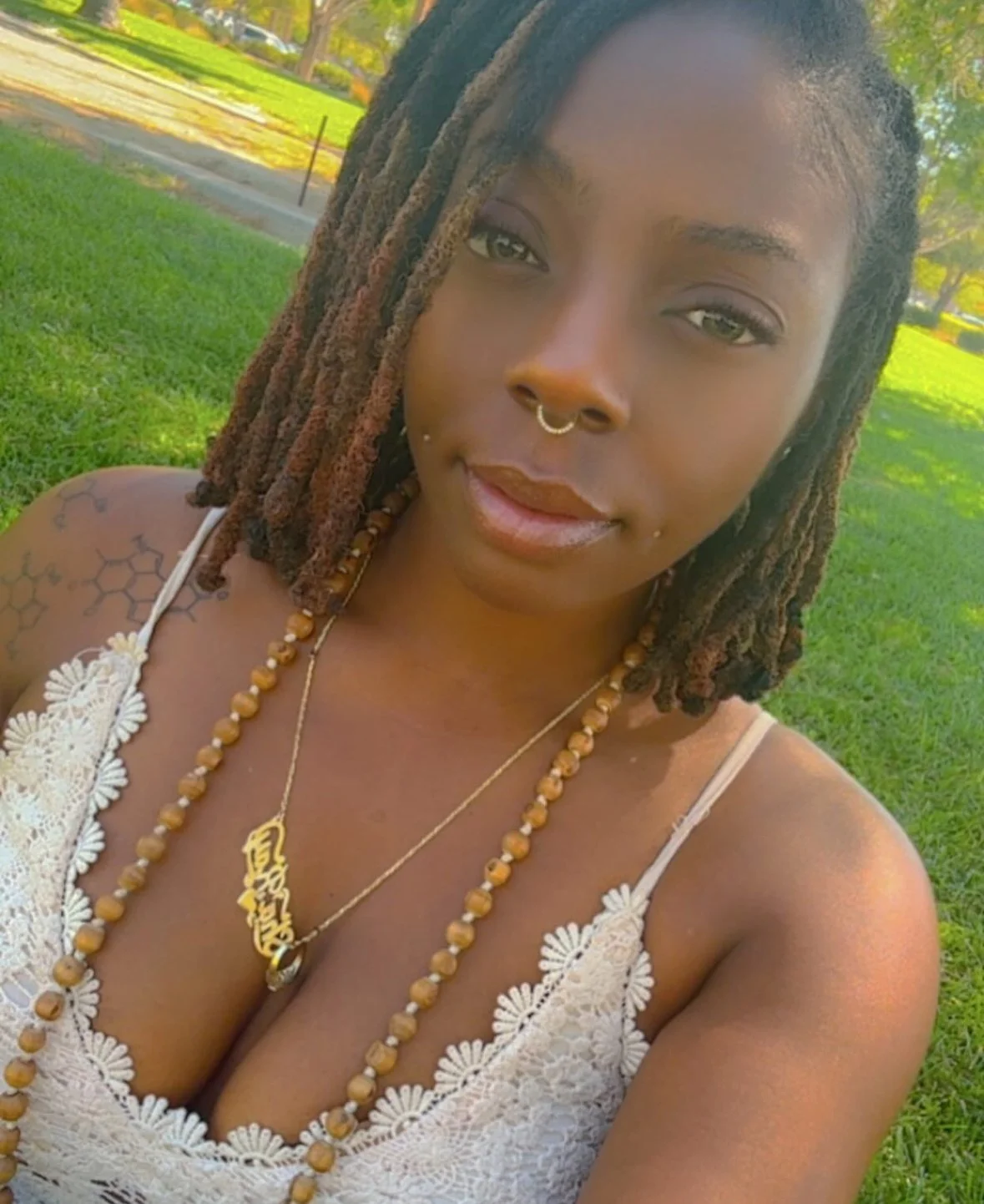 A woman with dreadlocks wearing a white lace top, gold nose and lip piercings, and beaded necklaces, standing outdoors on grass with trees in the background.