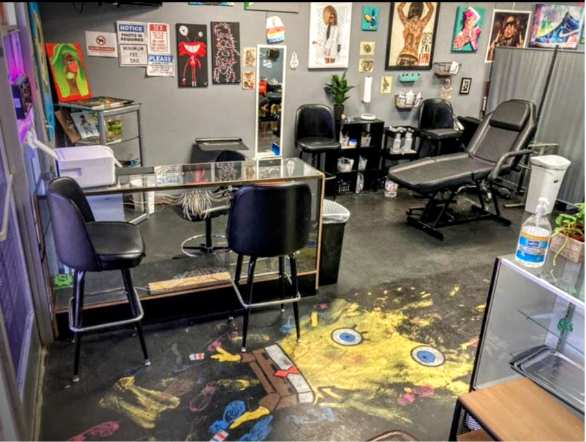 Tattoo studio interior with black chairs, artwork on the walls, a tattoo chair, and a colorful SpongeBob SquarePants-themed floor mat.