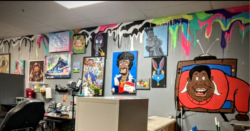 Colorful graffiti and vibrant paintings on a gray wall, including a cartoon man watching TV, a smiling cartoon character, and various abstract and pop art pieces in an artsy room.