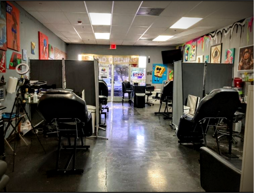 Tattoo studio with multiple tattoo chairs separated by temporary dividers, artwork on the walls, and a front door with an 'Open' sign visible outside.