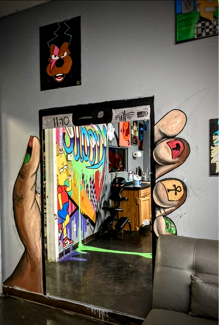 A mirror on a wall with cartoonish detailed hand-painted images around it, depicting fingers with rings. The mirror reflects a room with colorful graffiti-style art, a wooden cabinet, and a black massage or therapy chair. There are also framed cartoon artwork, including a character with an angry face, on the wall.