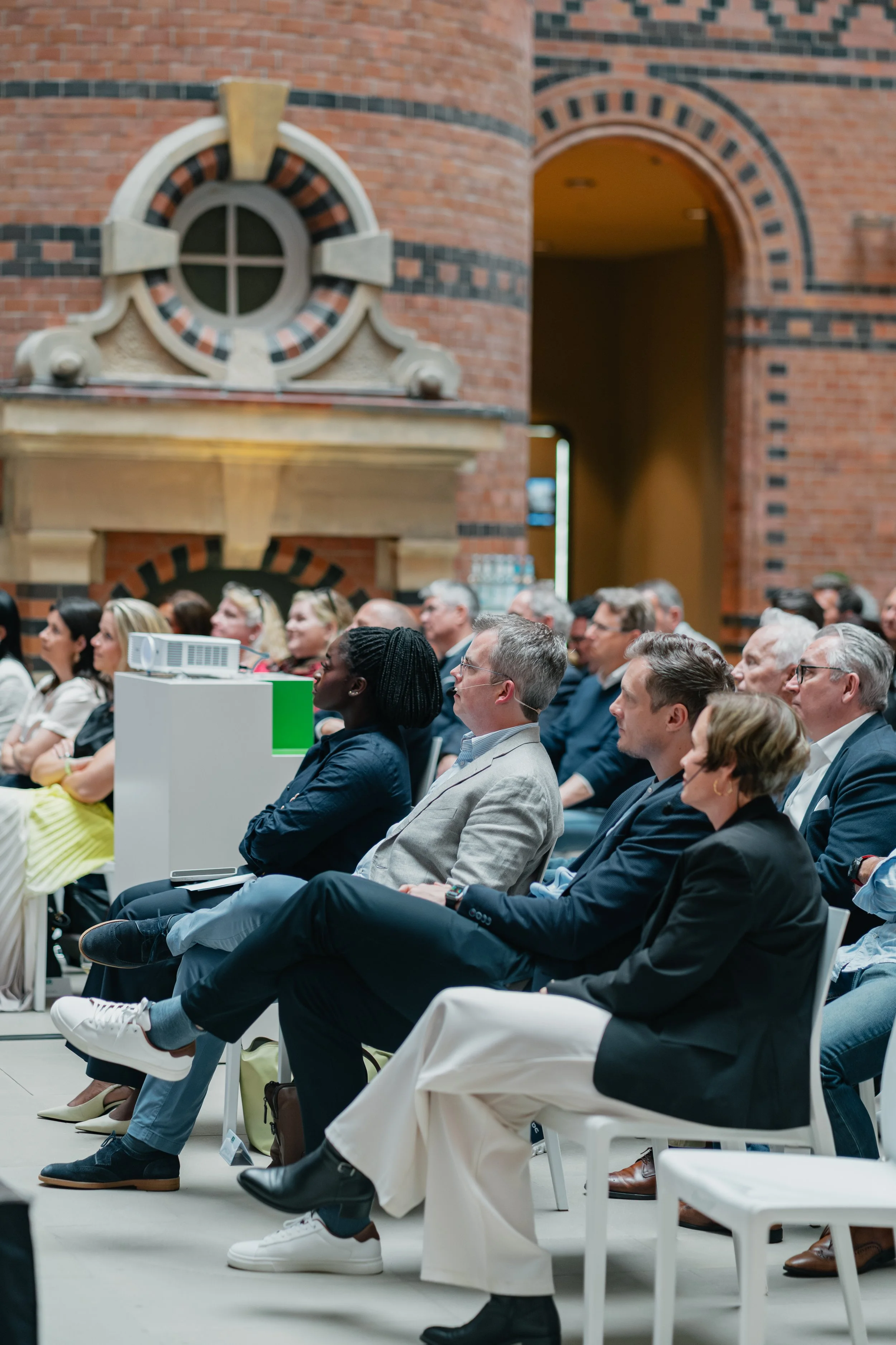 Audience seated in a conference room with brick walls, listening to a speaker, with a projector placed in front.