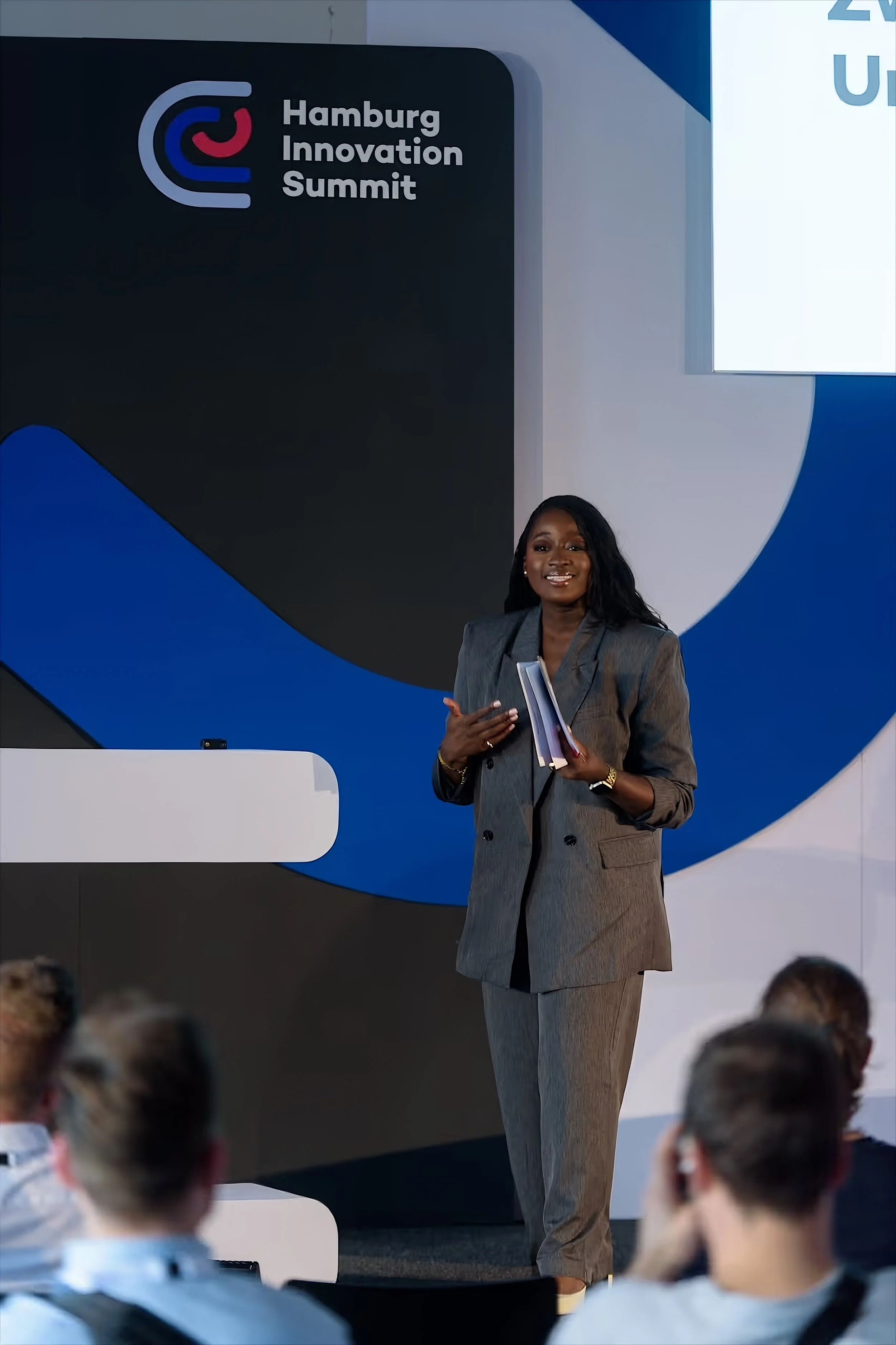 A woman speaking at the Hamburg Innovation Summit, standing on a stage with a blue and black background. She is holding notebooks, wearing a gray suit, and is addressing an audience.