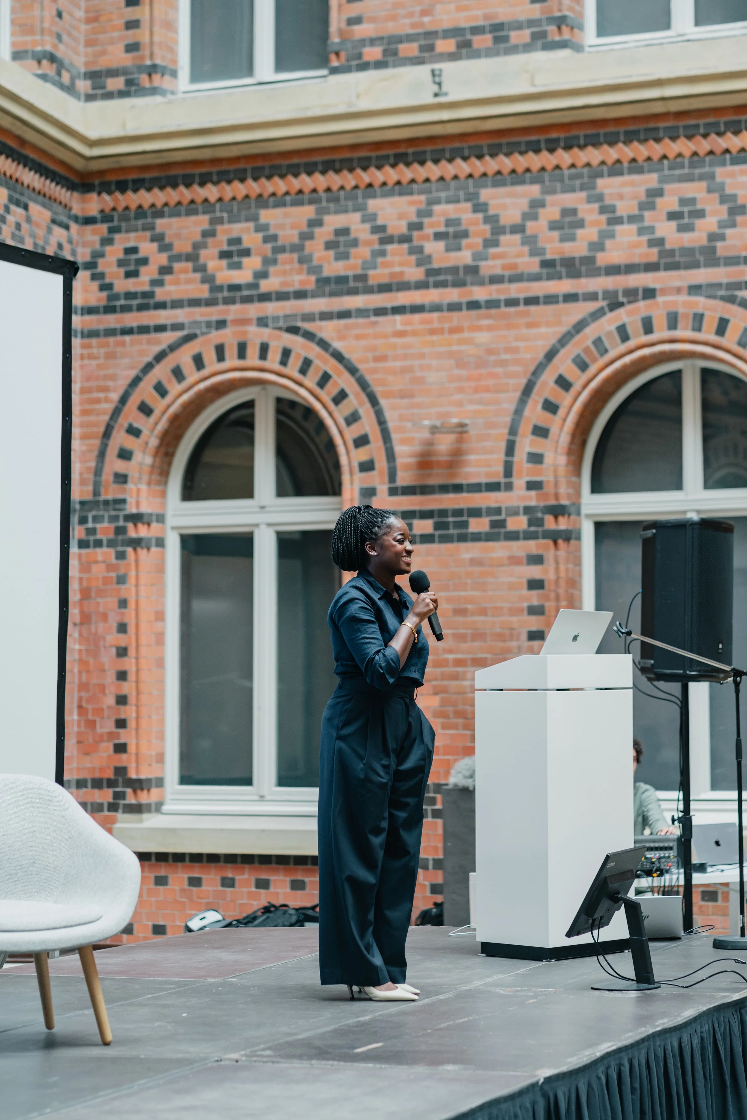 A woman in black pants and a dark blue blazer is speaking into a microphone on a stage outdoors against a brick building with arched windows. There is a white podium with a MacBook on it and a sound system nearby.