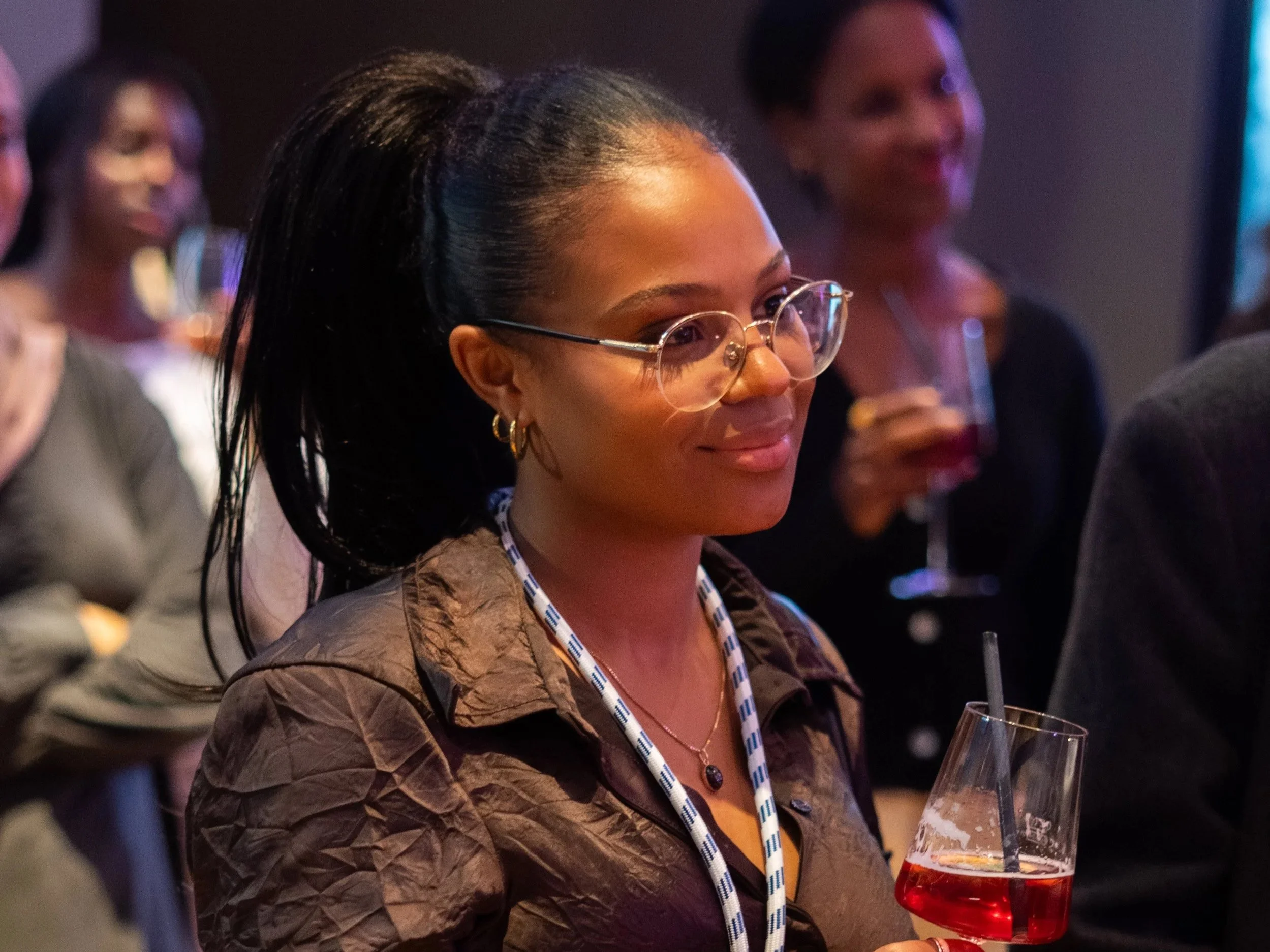 Young woman with glasses and earrings, smiling and holding a drink with a straw at what appears to be a social event or gathering.