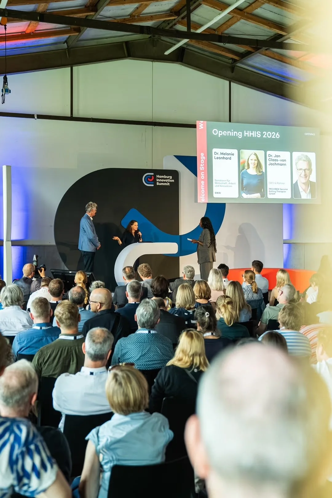 People attending the Hamburg Innovation Summit in a large conference hall. On stage, three individuals are engaged in a presentation, with a large screen displaying names and titles of speakers, Dr. Melanie Leonhard and Dr. Jan Claas-von Jachmann. The audience is seated, listening attentively.