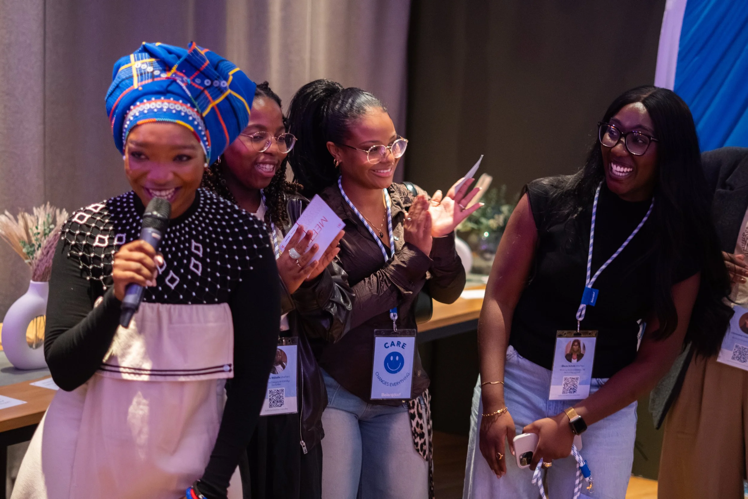 Group of women standing together at an event, smiling and engaging in conversation. One woman is wearing a colorful headwrap, holding a microphone, and the others are wearing eyeglasses and conference badges.