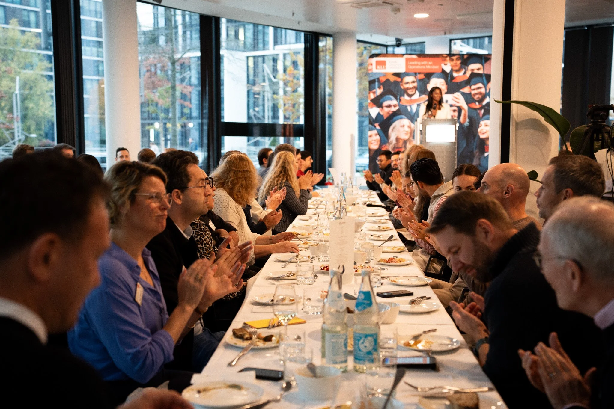 People sitting at a long banquet table in a modern conference room, clapping during a graduation celebration, with a large banner of graduates in caps and gowns in the background.