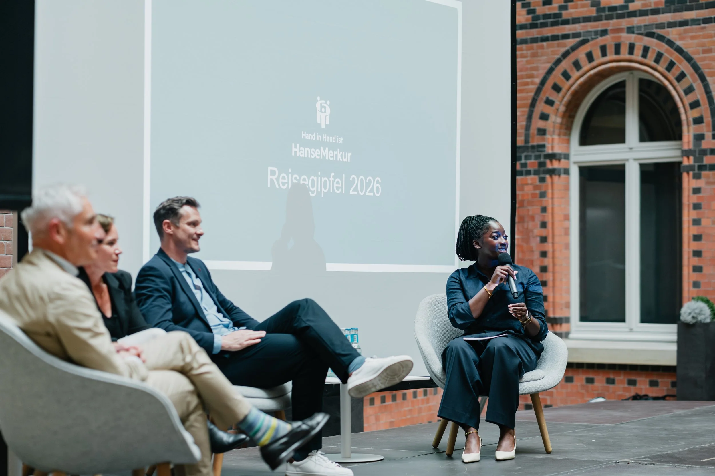 Panel discussion with four people seated on stage, with a woman holding a microphone speaking, and a large screen behind them displaying the title "Hand in Hand ist HaneMerkur Reigepitel 2026". The setting appears to be a formal outdoor event in front of a brick building.
