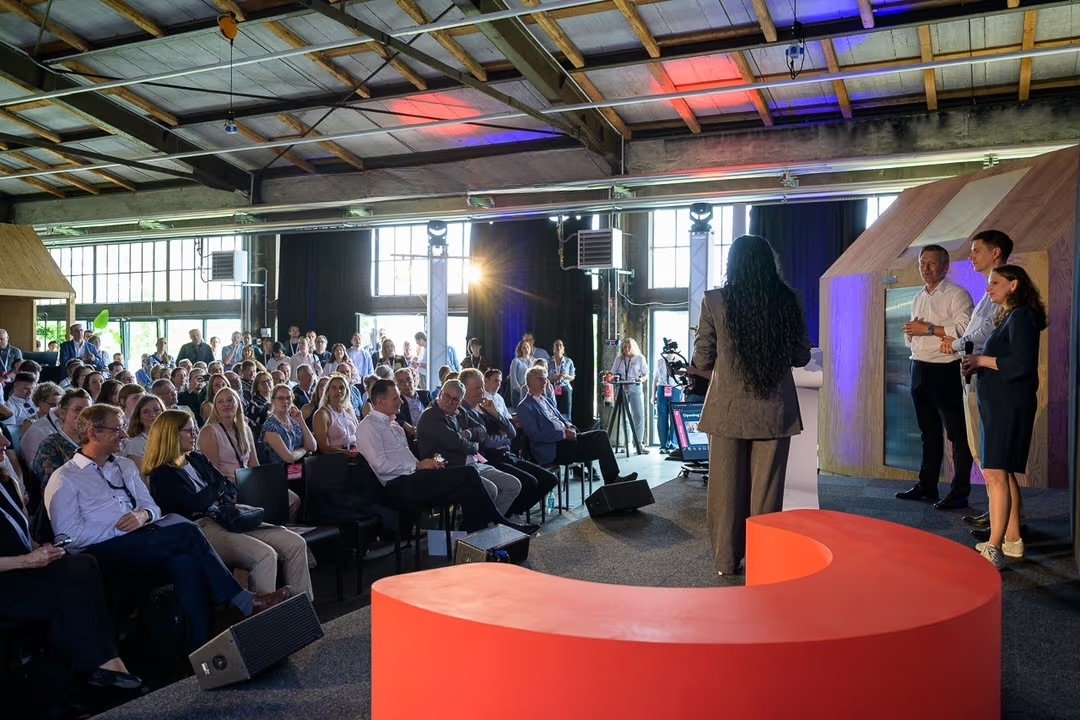 A woman giving a presentation on stage to an audience in a large industrial-style space with exposed beams and large windows, with three other people on stage and a camera operator recording the event.