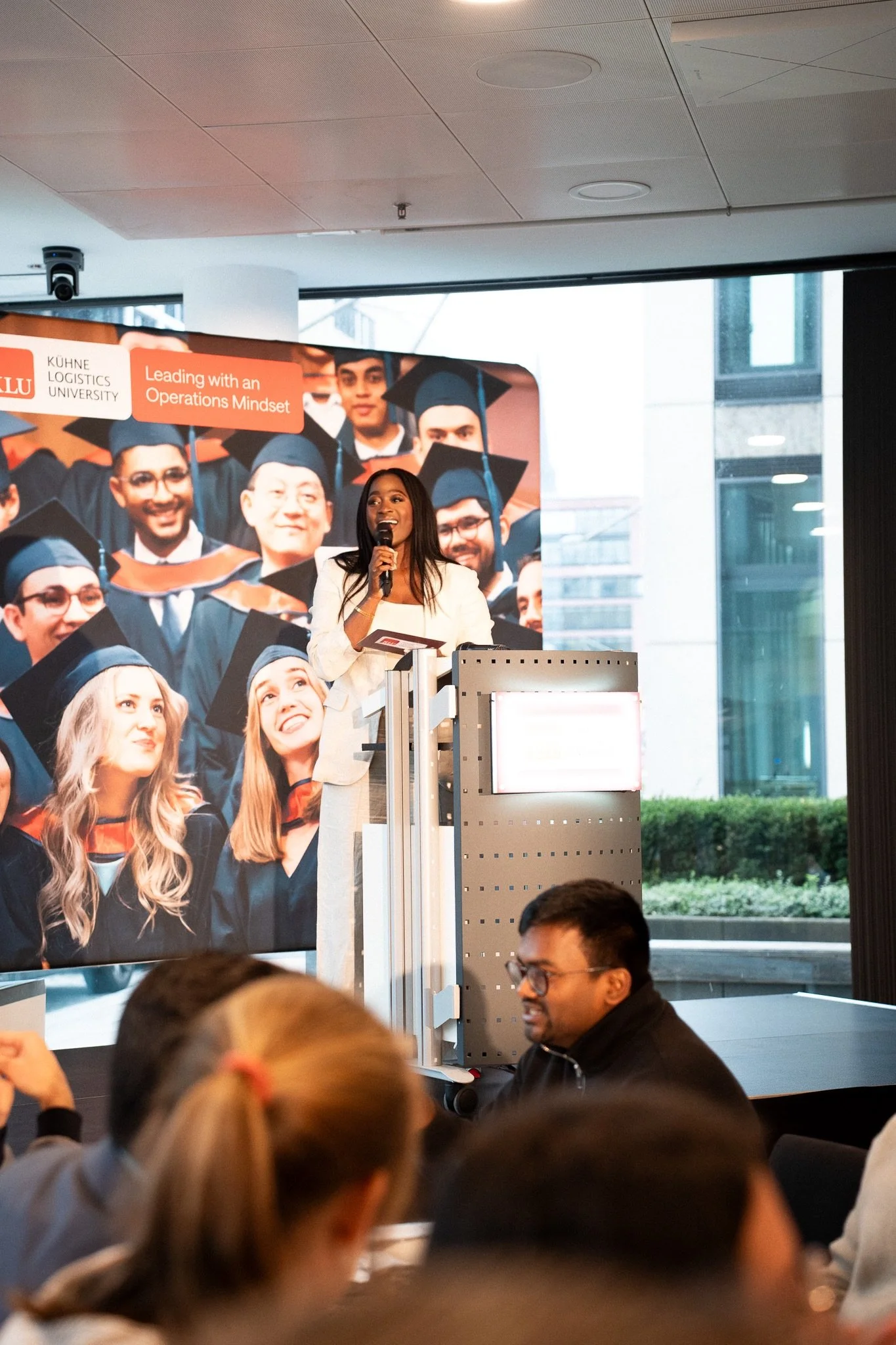 A woman standing behind a podium, speaking into a microphone at an event. In the background, there is a large poster depicting several students in graduation caps and gowns, with text about a university leading with an operations mindset. The setting appears to be a modern conference or university event space with large windows.