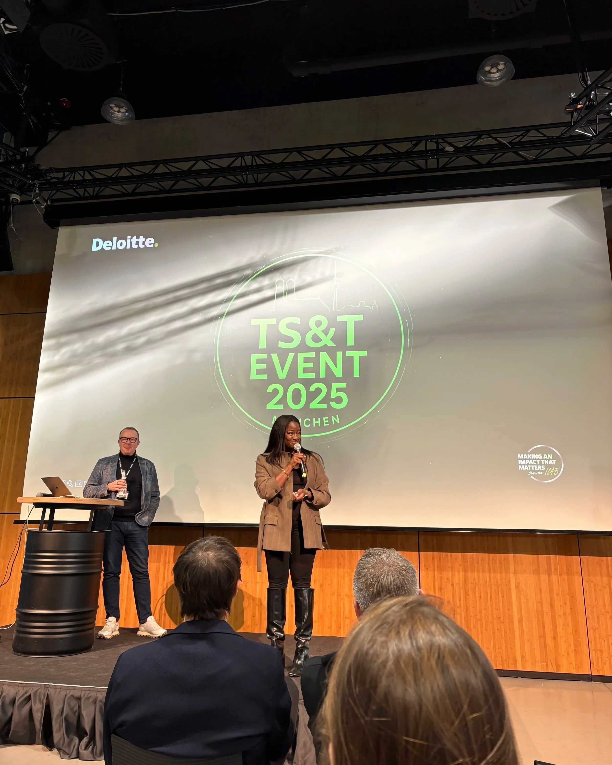 A woman is speaking into a microphone on a stage at the TS&T Event 2025 in Munich, with a man standing nearby. The large screen behind them displays the event details and logo, along with the Deloitte logo at the top left corner.