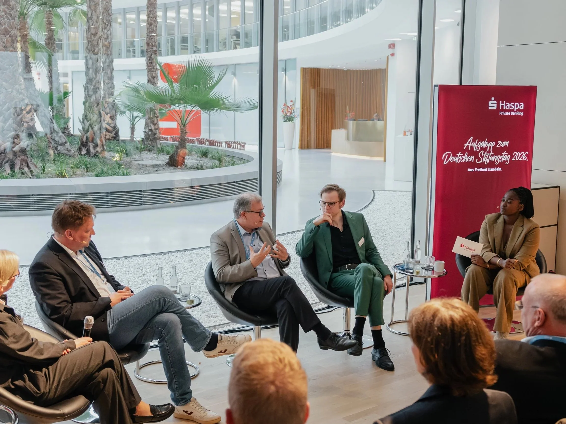 Business professionals engaged in a panel discussion at a conference, seated in a semi-circle in a modern building with large windows, indoor plants, and a red banner with white text.
