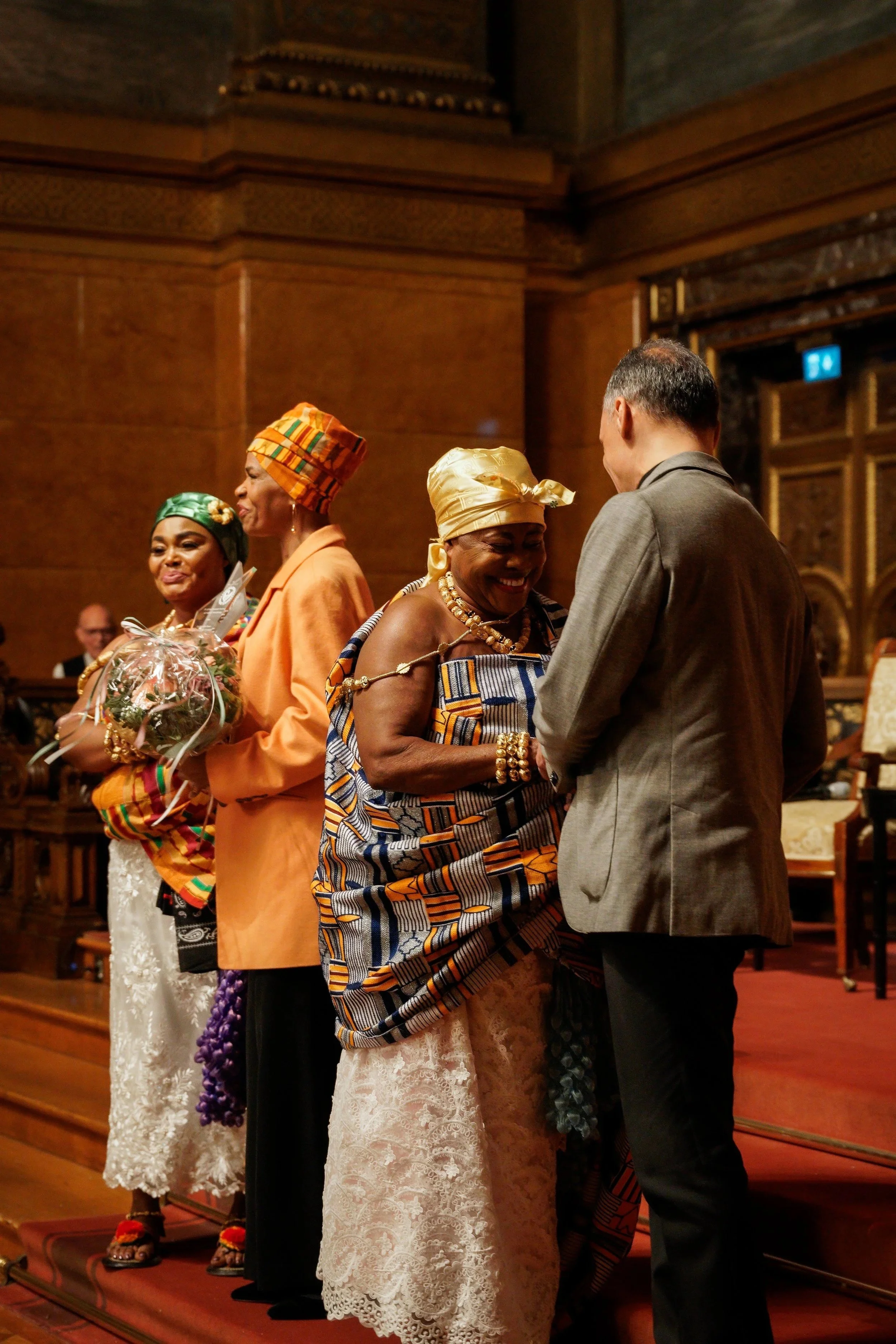 People dressed in traditional African attire and a man in a gray suit talking and sharing a smile in a formal setting with wooden paneling and ornate decor.