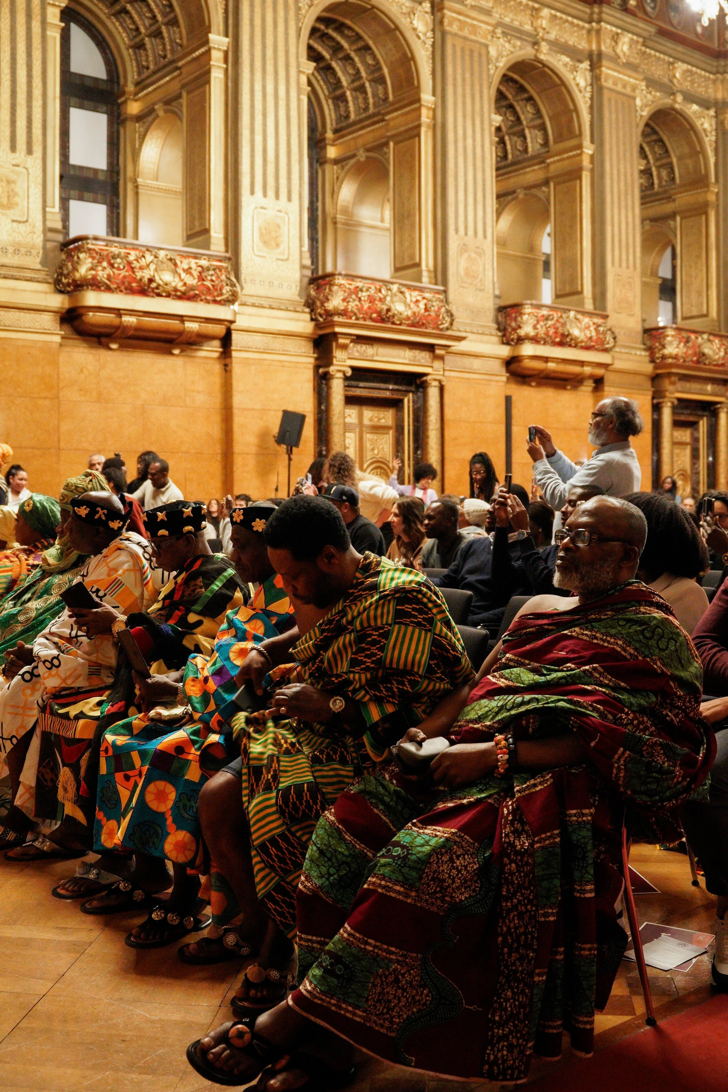 A diverse group of people seated in a grand, ornate hall with gold-trimmed walls, detailed arches, and decorative balconies, attending an event.