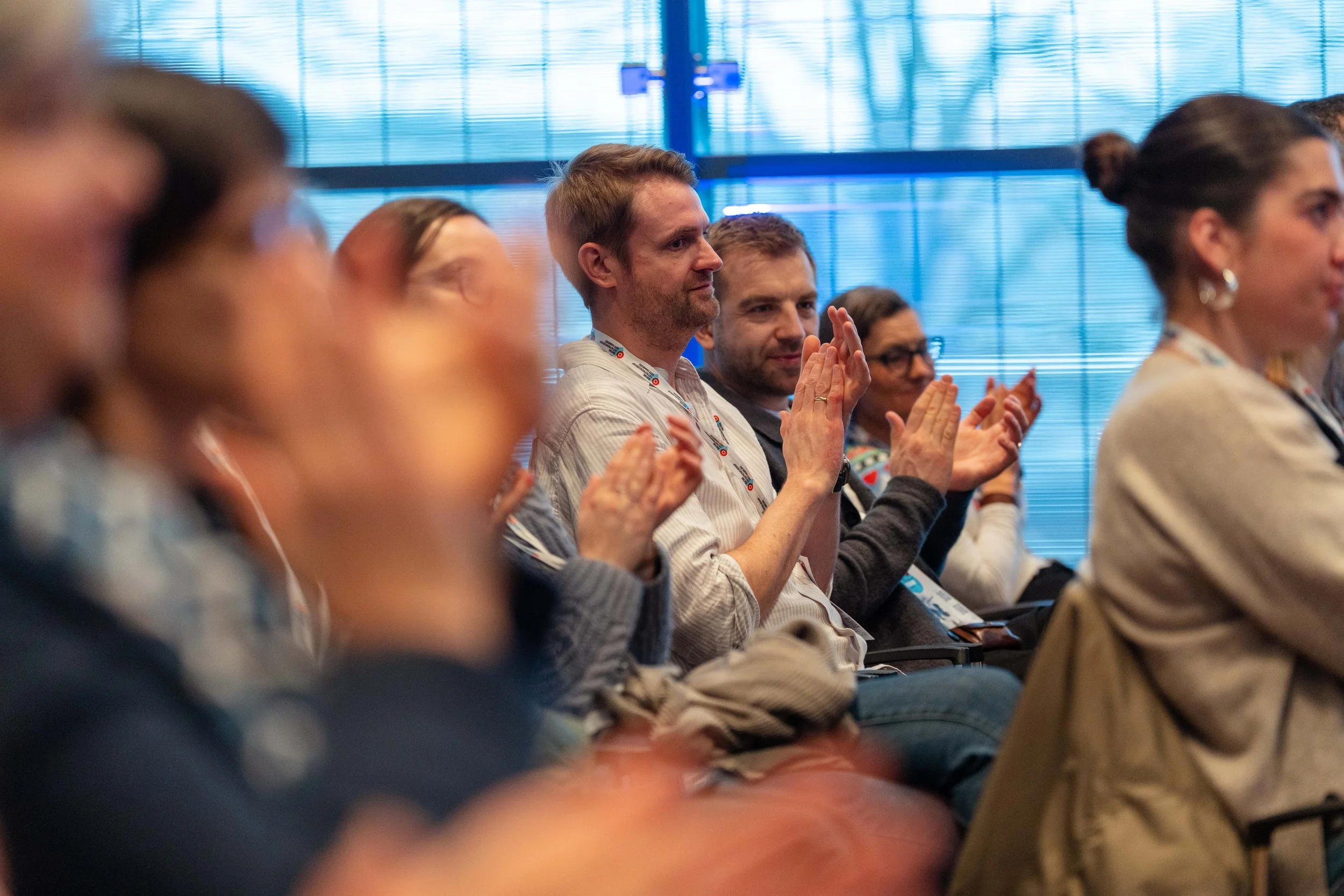 People attending a conference or seminar, sitting in rows and clapping. Focus on a man with light brown hair and a beard in the middle. The background includes large windows with blinds.