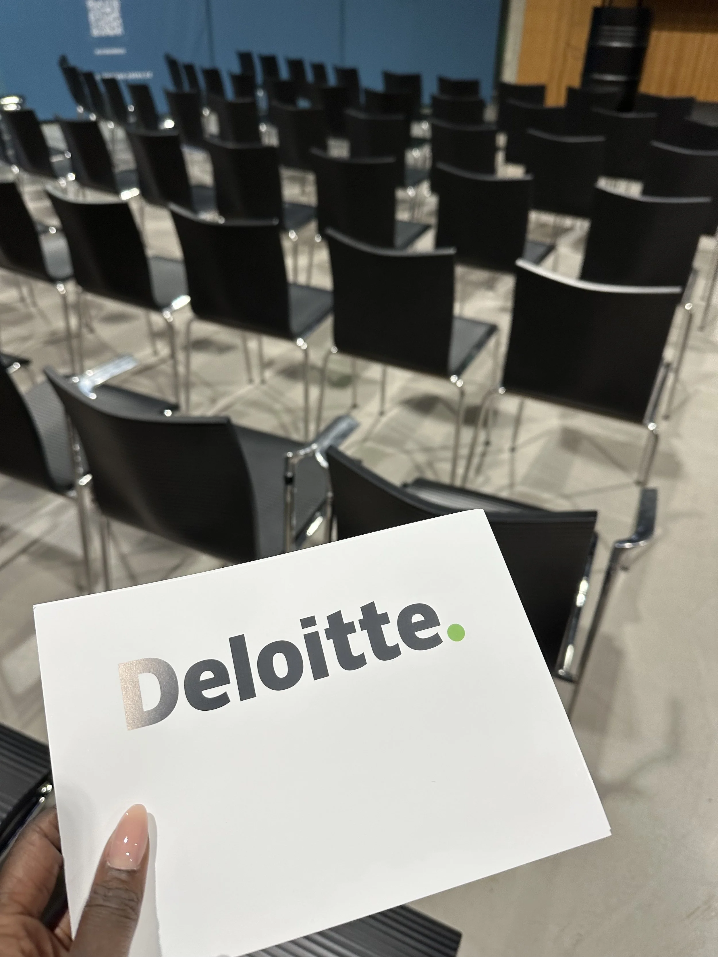 Person holding a white paper with the Deloitte logo printed on it, in a room with multiple empty black chairs arranged in rows.