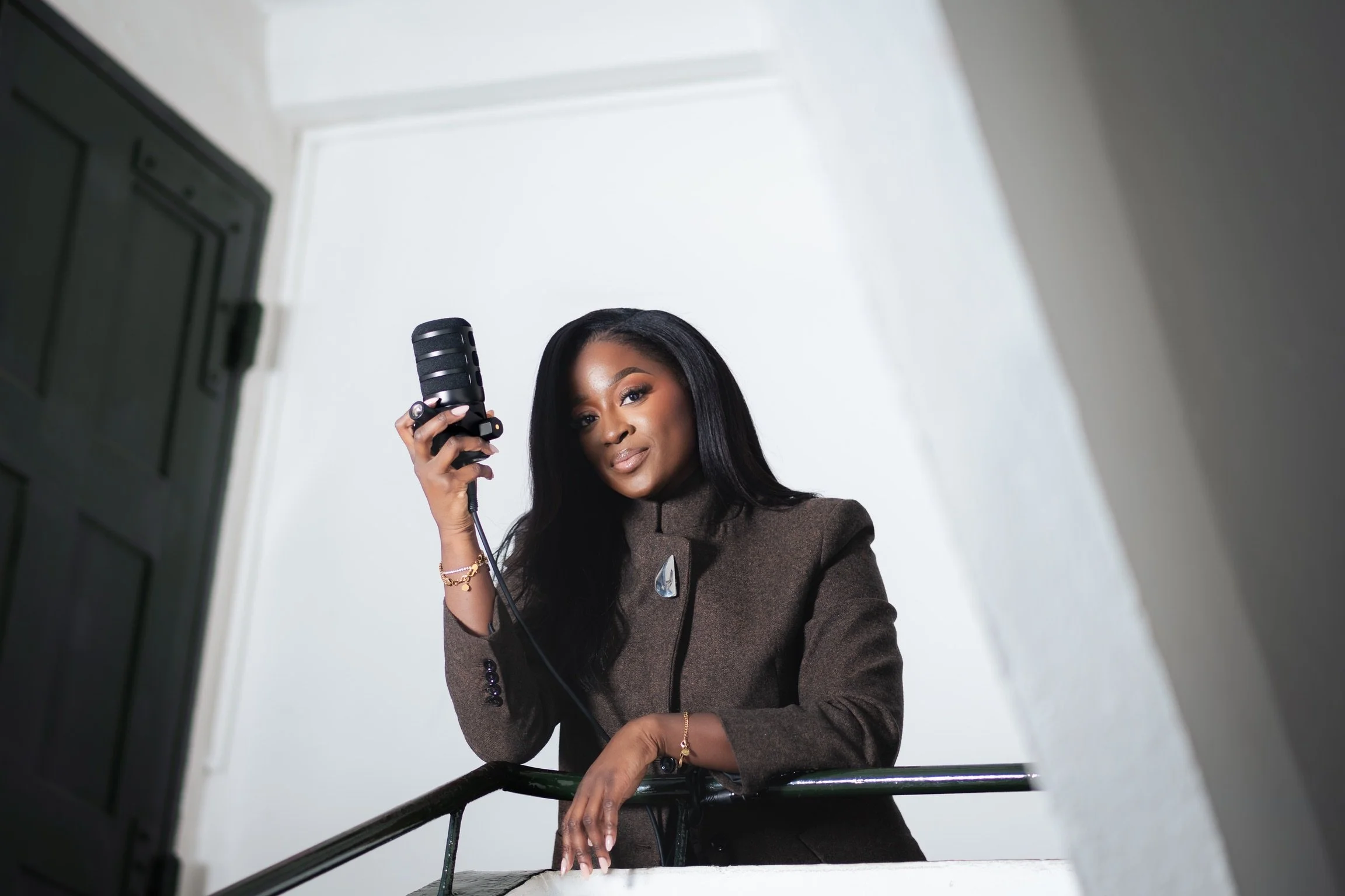 A woman with long black hair and brown skin examining herself in a mirror with a hairdryer in hand, standing in a well-lit room with white walls and a black railing.