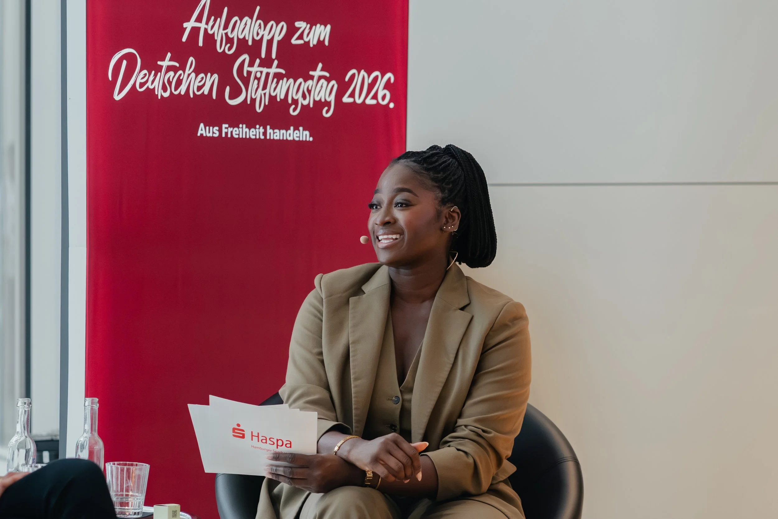 A woman with dark skin, braided hair, wearing a beige blazer, smiling and sitting at a table with a red banner behind her that has white text in German.