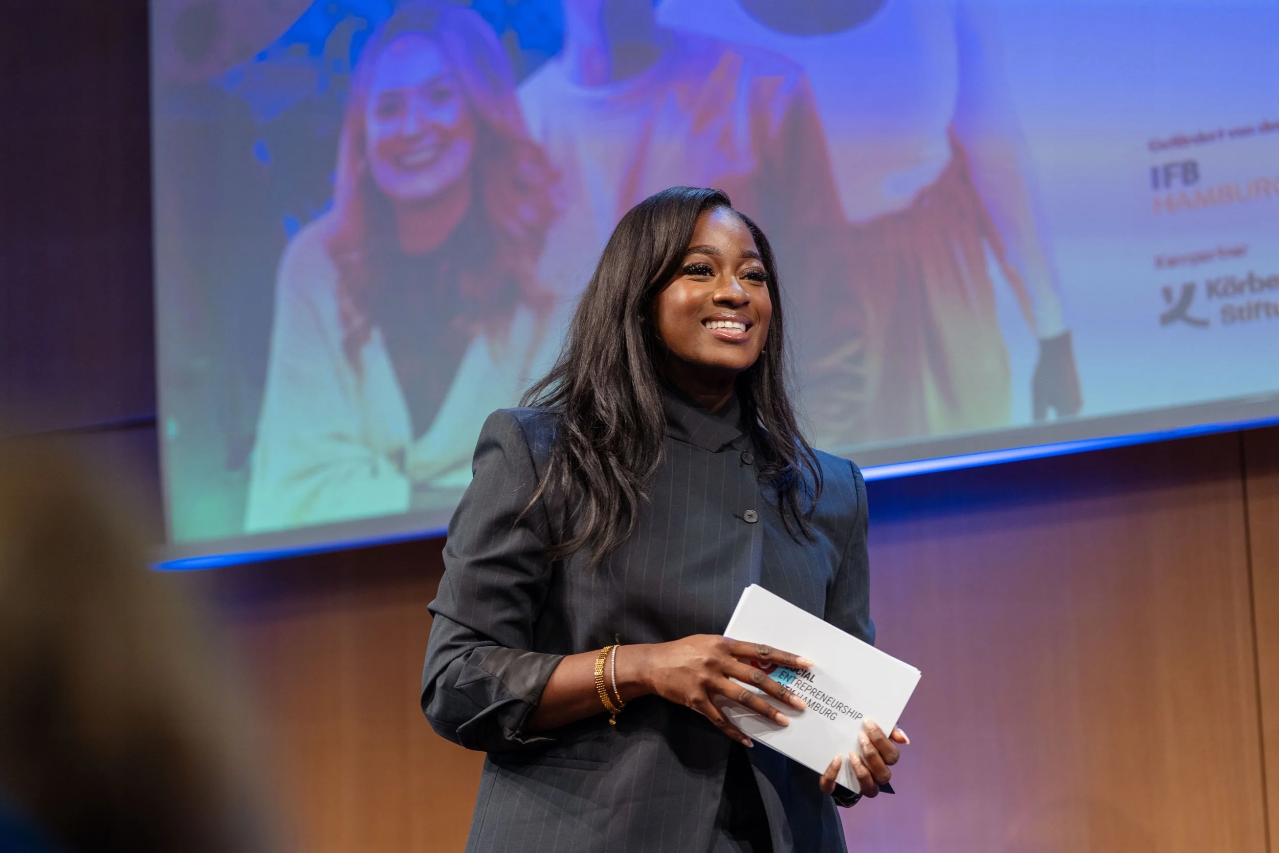 A woman with long black hair, dressed in a dark pinstripe suit, holding a white booklet with the title 'Entrepreneurship' and a pen, smiling during a presentation, with a large projection screen behind her showing a similar image of her.