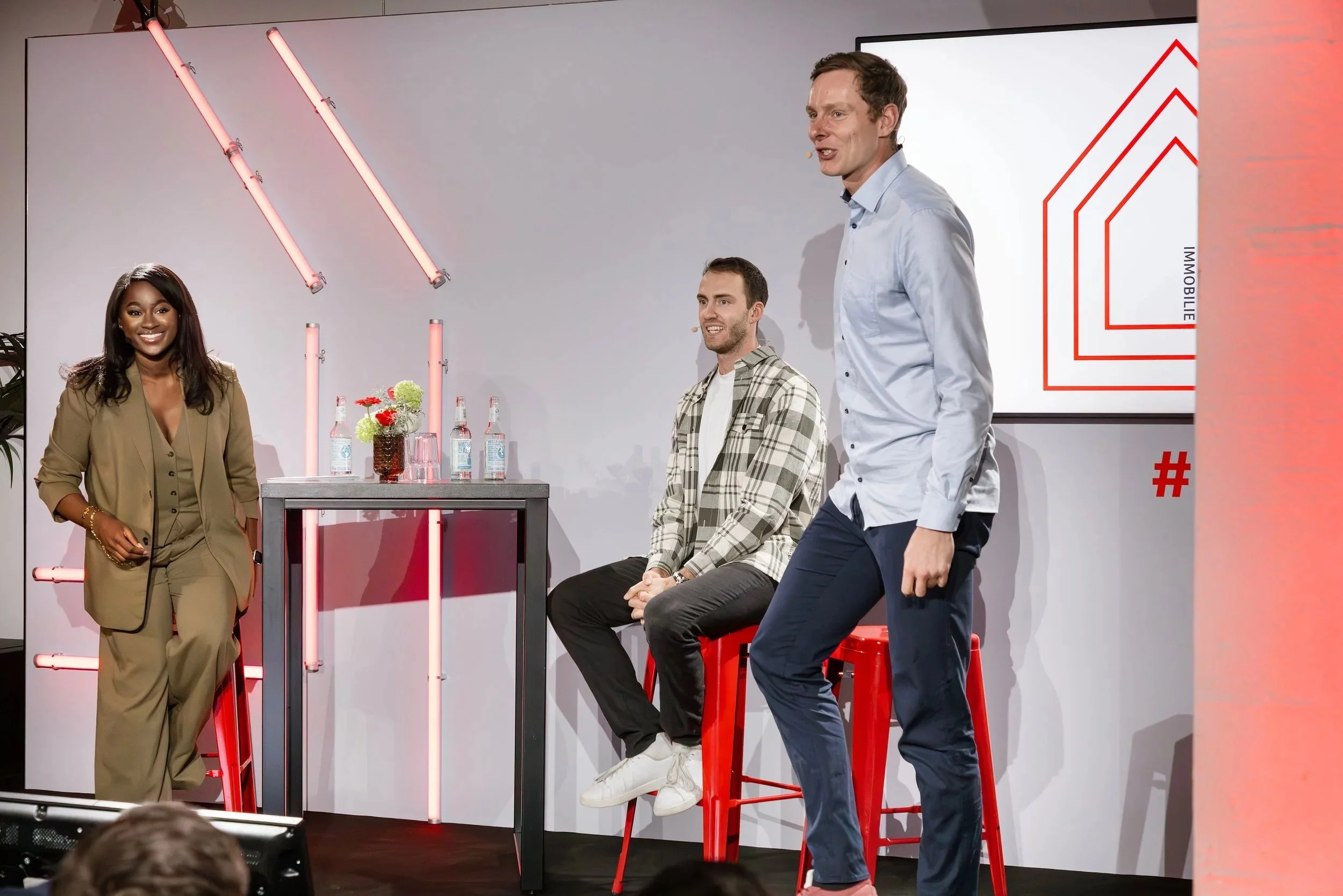 Three individuals on a stage with a modern backdrop. One woman in a tan suit is standing, smiling, and wearing gold jewelry. One man with a beard, in a checked shirt, is sitting on a high red stool. The third man, in a light blue shirt, is standing and speaking. There are decorative pink lights, bottles of water, and a vase with flowers on a small table.