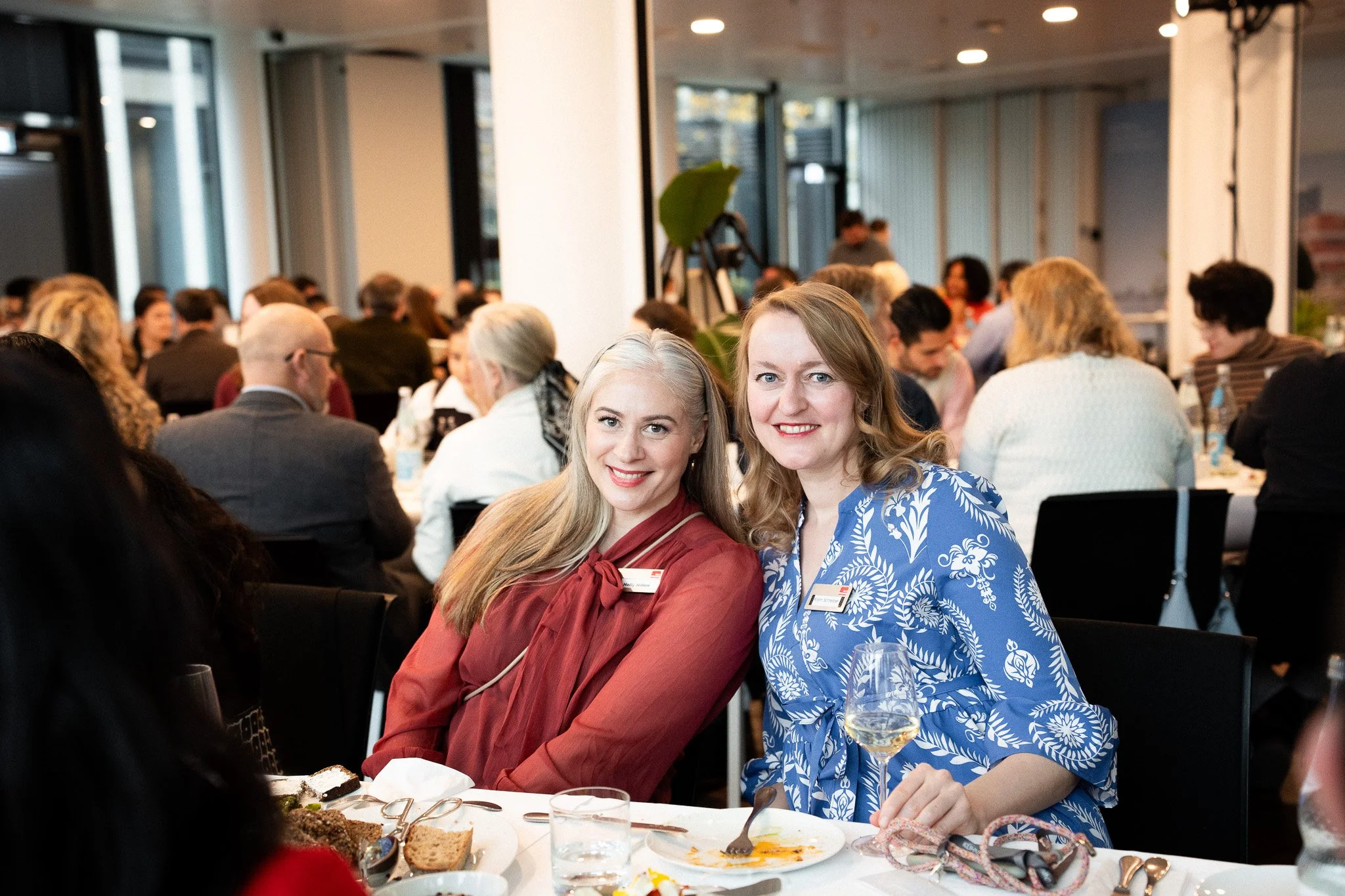 Two women smiling at camera while seated at a table during a gathering or event, with many people seated and mingling in the background, and food and drinks on the table in front of them.