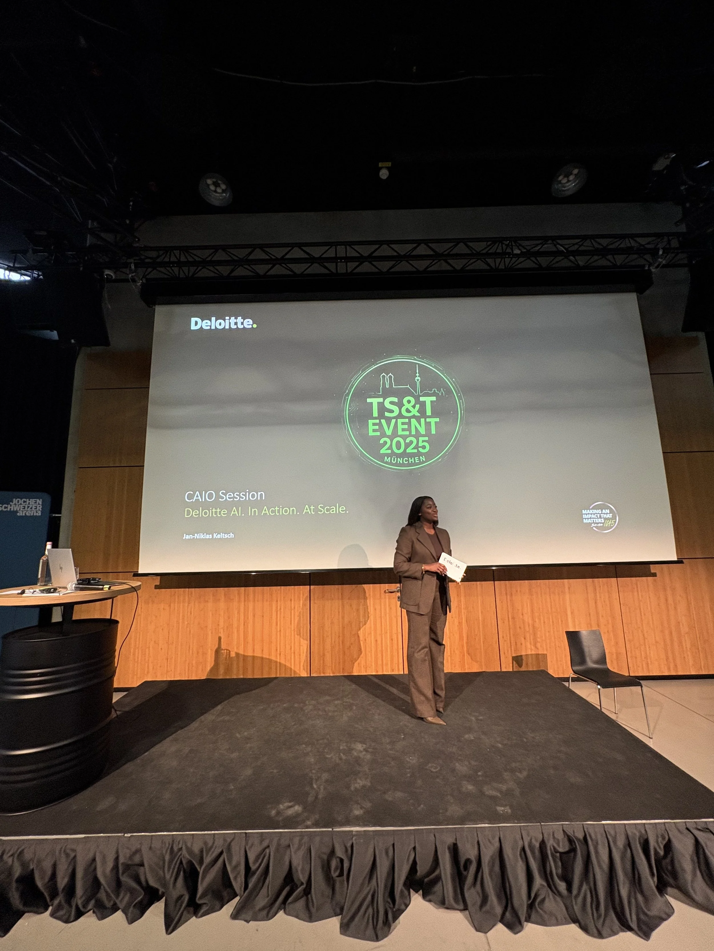 A woman standing on a stage in front of a large screen at the TS&T Event 2025 in Munich, delivering a presentation about Deloitte AI. In Action. At Scale.