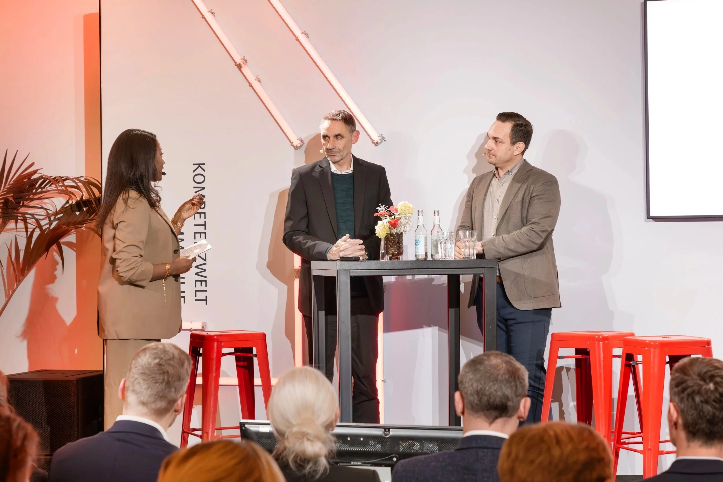 Three people stand behind a high table on a stage, engaging in a discussion during a panel or presentation. The woman on the left is holding some notes, the man in the middle is looking on, and the man on the right is listening. The audience is seated facing the stage. There are three red stools in front of the table, a small bouquet of flowers and beverage bottles on the table, and a large blank screen on the right side of the stage. A large illuminated light fixture is mounted on the white wall behind them.