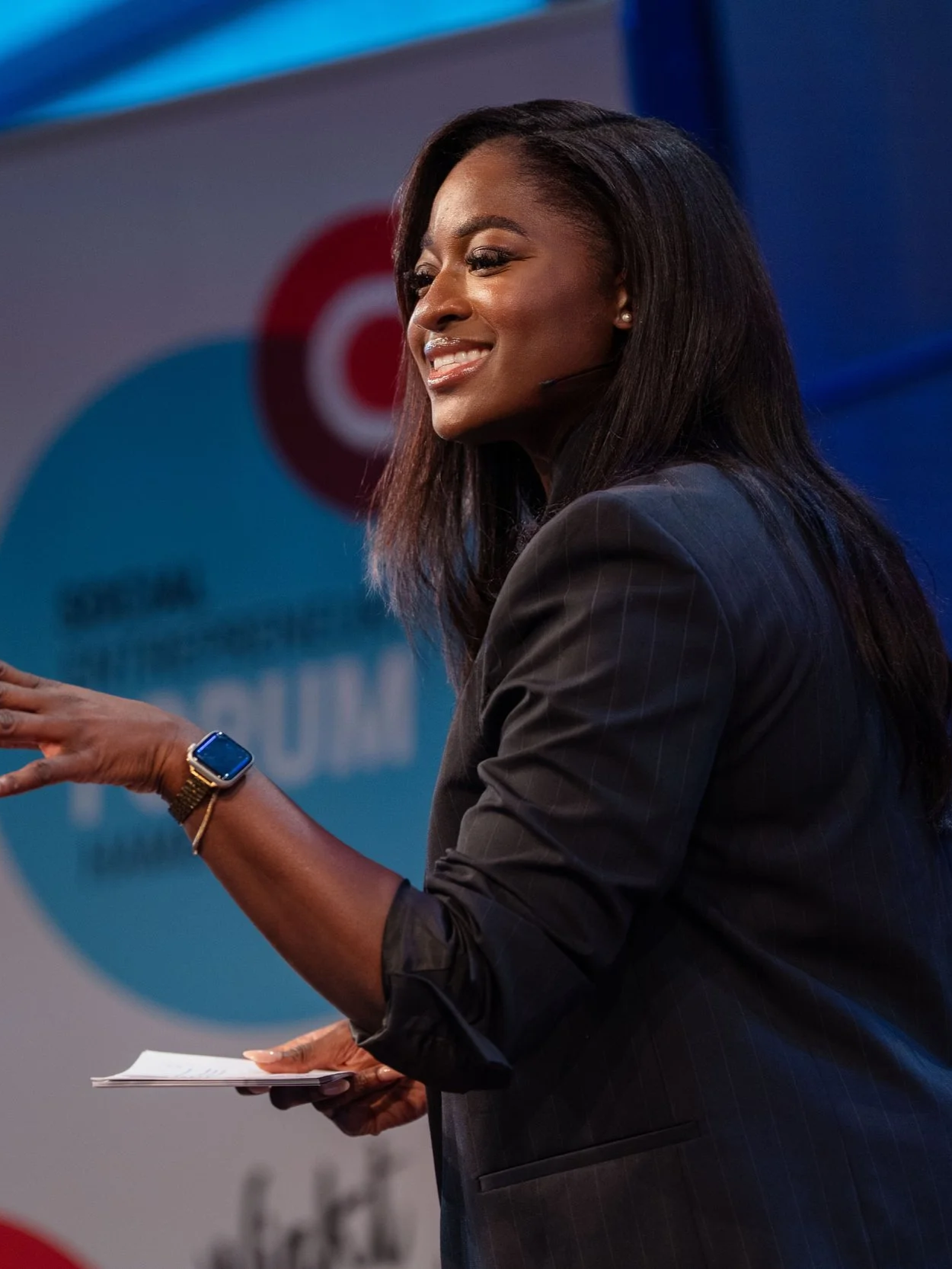 A woman in professional attire speaking at a conference, holding notes, with a background featuring a Target logo and a blurred audience.
